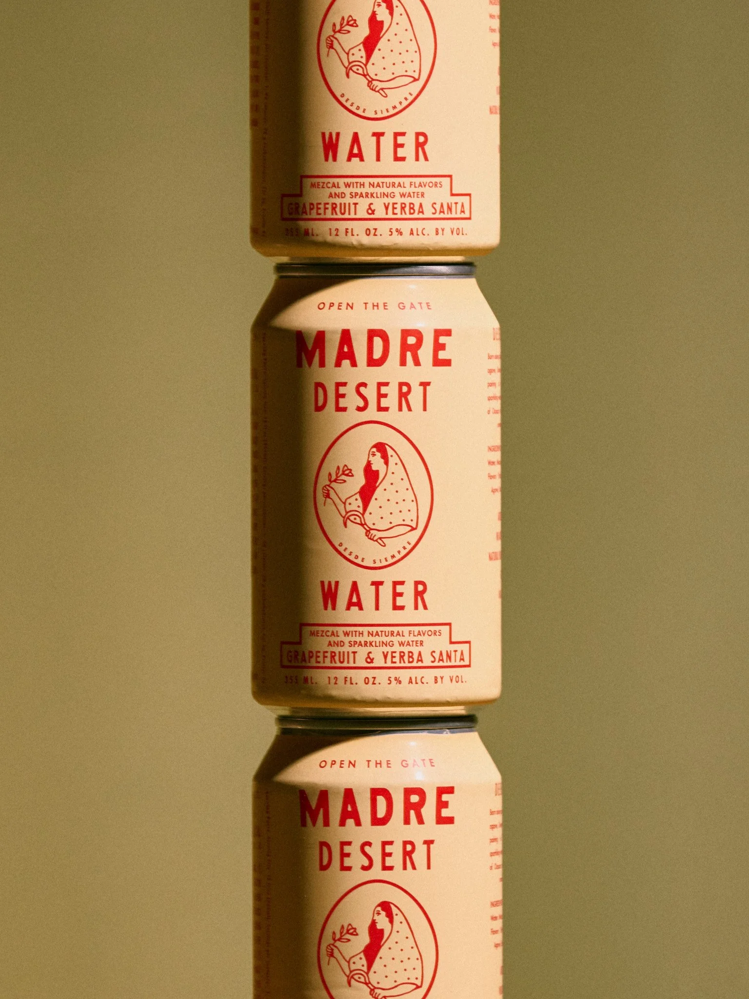 Three cans of Madre Desert Water, stacked vertically, with a label featuring a woman holding a plant, in red and white. The cans are reflected on a surface and are set against a plain background.