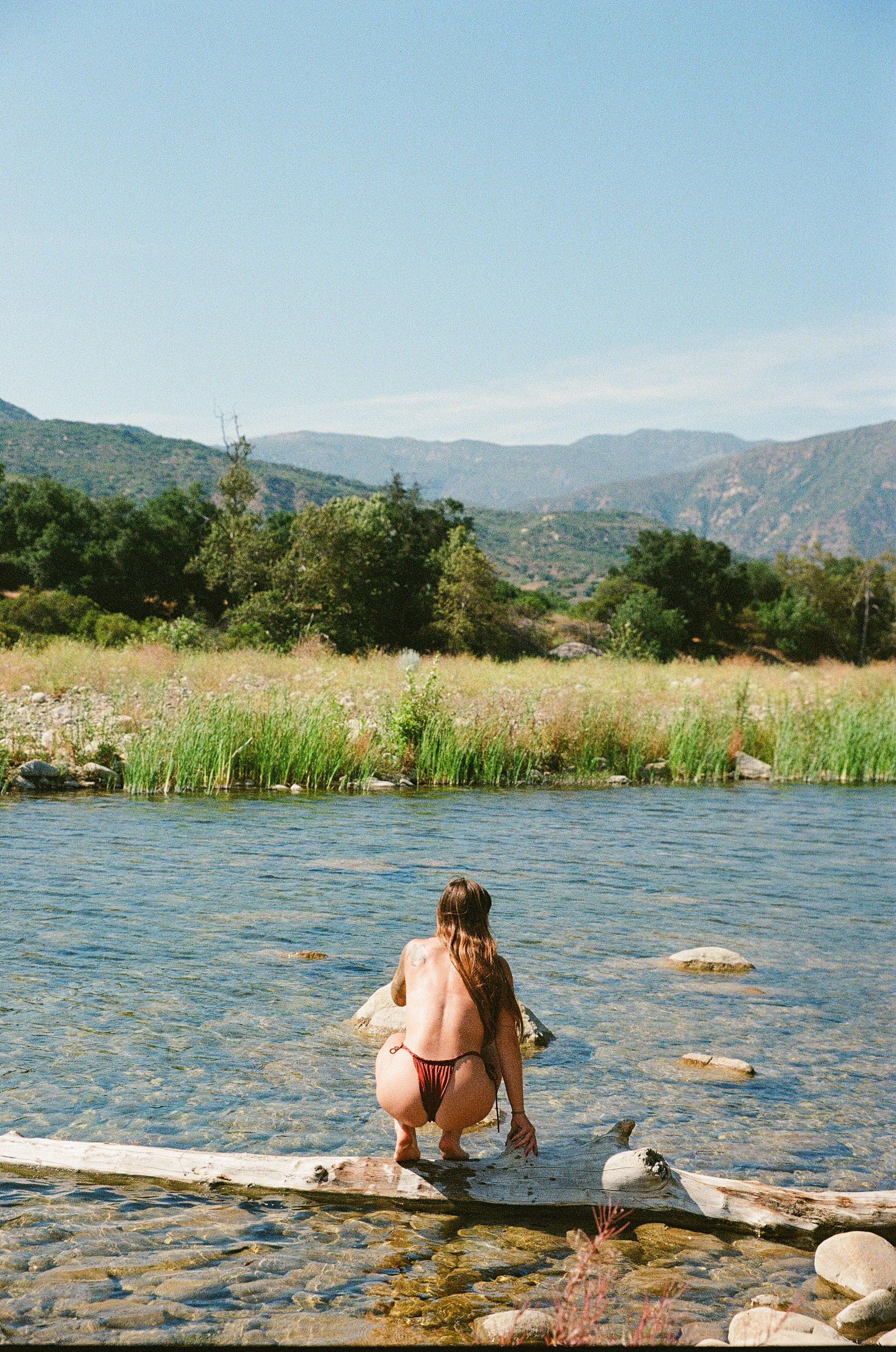 A woman with long hair squatting on a log in a shallow river, with mountains and trees in the background under a clear blue sky.