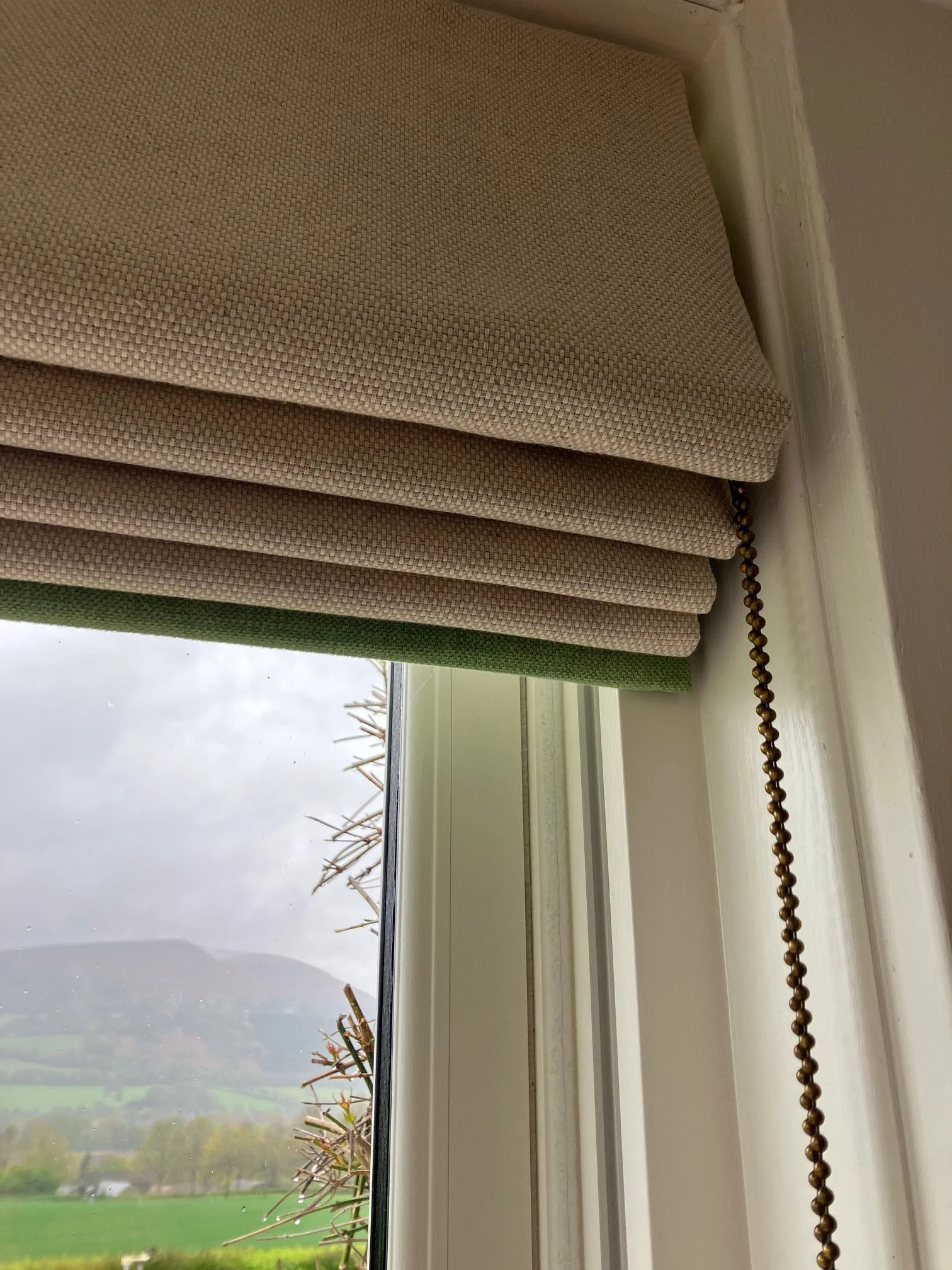 A close-up view of a beige linen roman blind with green trim and antique brass chain, revealing a foggy, rainy landscape with plants and open fields outside.