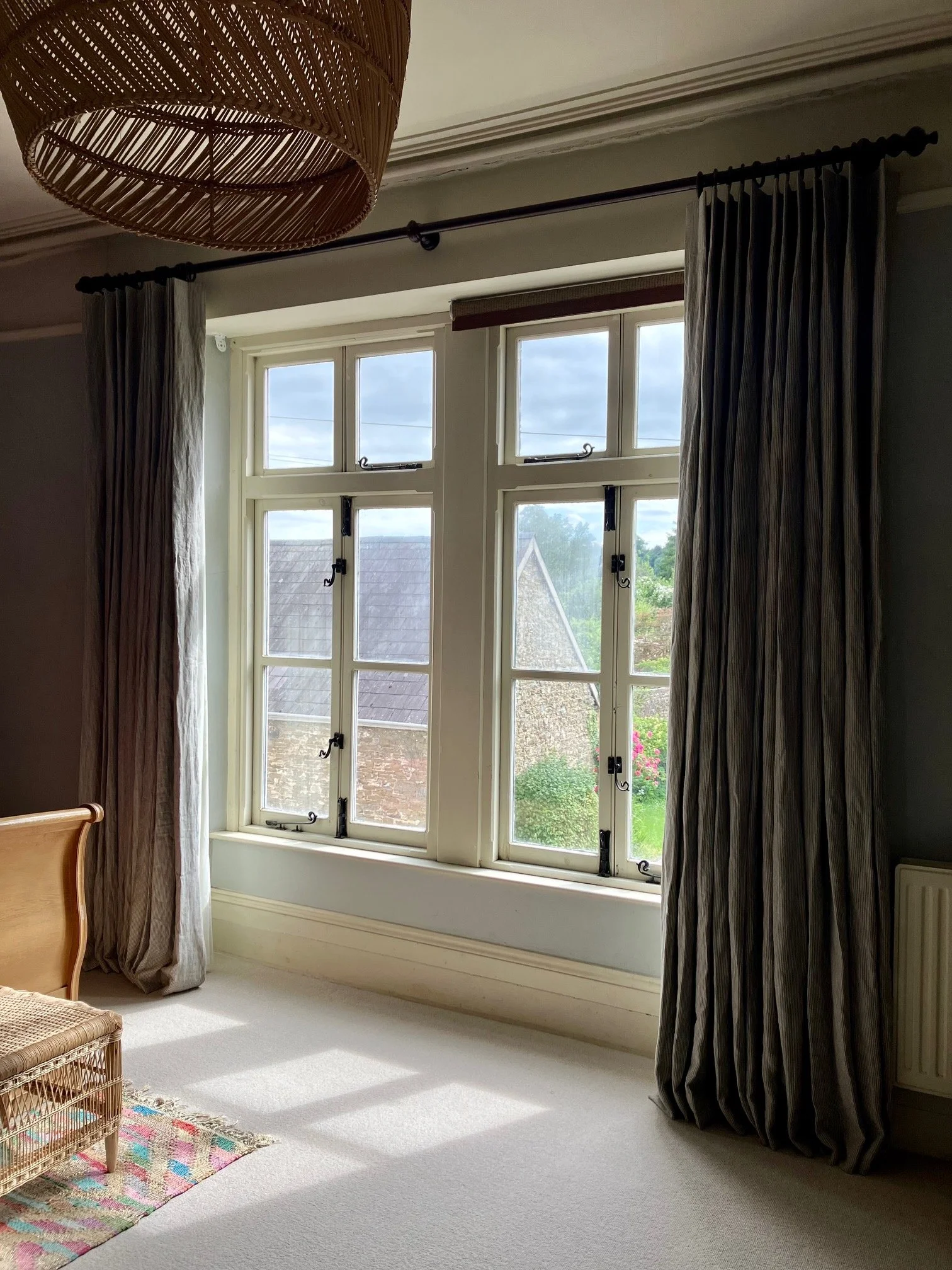 View of a bedroom with large victorian window, striped curtains, and a wicker chair beside a colorful patterned rug and cream carpet.