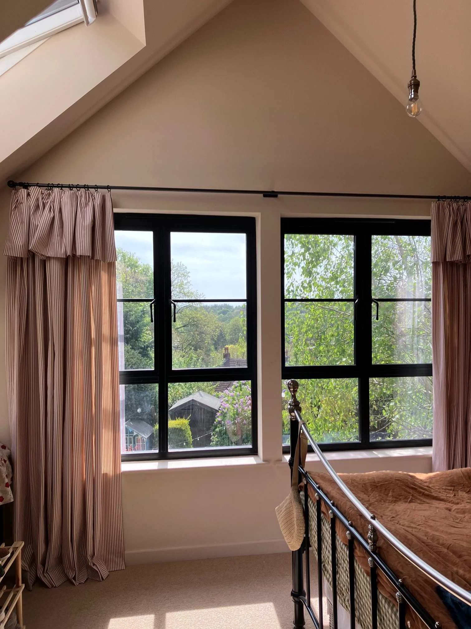 View of a bedroom with large black-framed windows, pink and white striped curtains, and a bed with a brown floral blanket. Outside the windows, there are trees and a garden. A black metal bed frame is partially visible.
