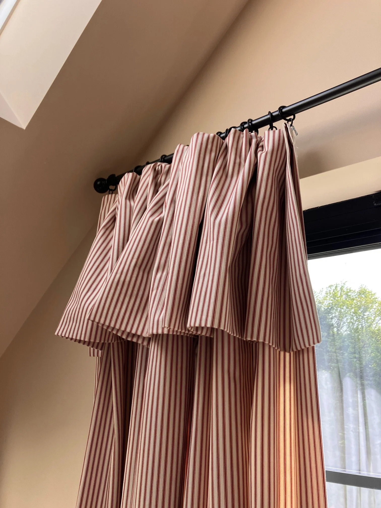 Red and white striped curtains with flop-over header hanging on a black steel curtain pole  in front of a window, partly opened to show some greenery outside.