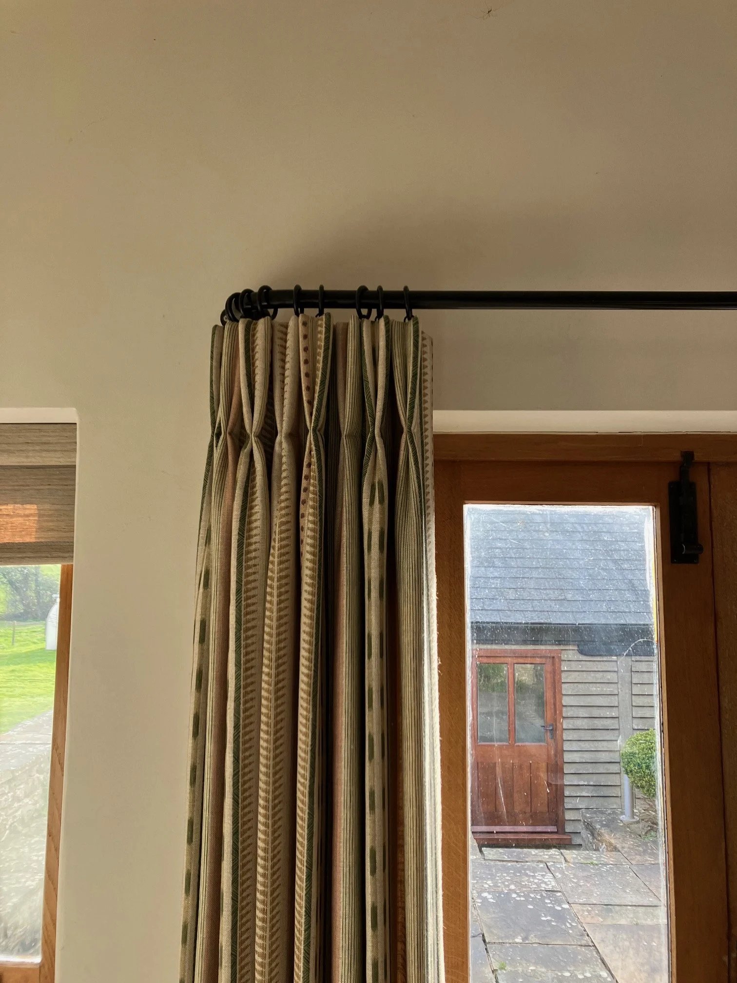 Close up of pattered, linen, interlined curtains, double pinch pleat header, hang from black metal french pole, showing an outdoor view of a garden or yard with a wooden shed and wet ground.