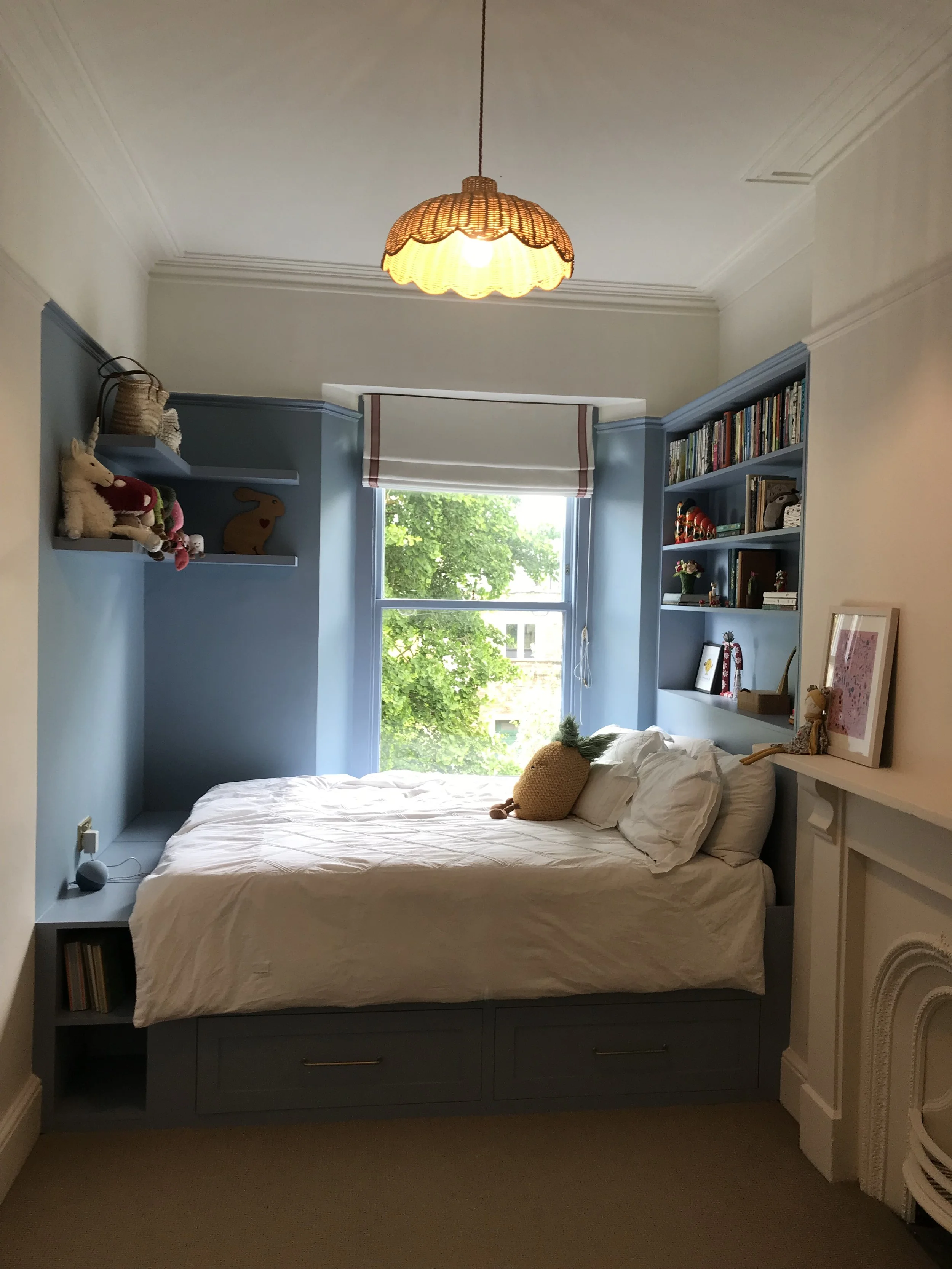 A bedroom with a cream Roman blind with pink trim, bed under a window, blue built-in shelves with books and stuffed animals, a framed picture on the nightstand, and a hanging light fixture with a woven lamp shade.