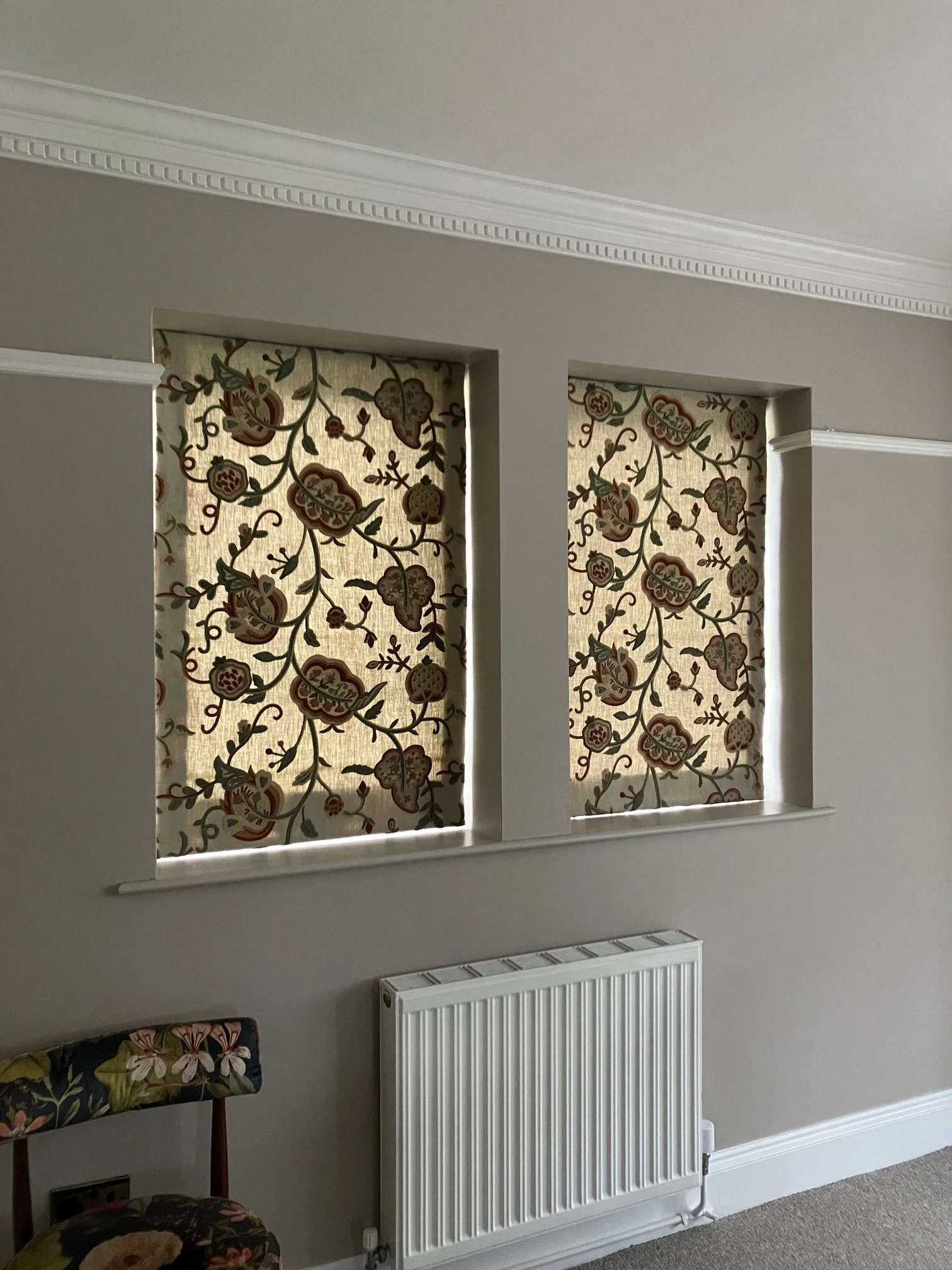 Interior view of a room featuring two windows with embroidered, patterned Roman blinds, a white radiator below the windows, and an upholstered chair with floral fabric in the lower left corner.