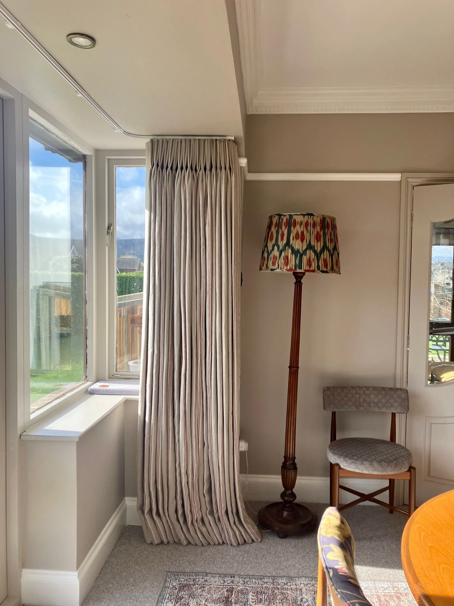 Striped interlined curtains with pinch pleat header hanging on ceiling track, a chair with a patterned cushion, and part of a round wooden table with a floral chair. The room has beige walls, white trim, and a grey carpet.