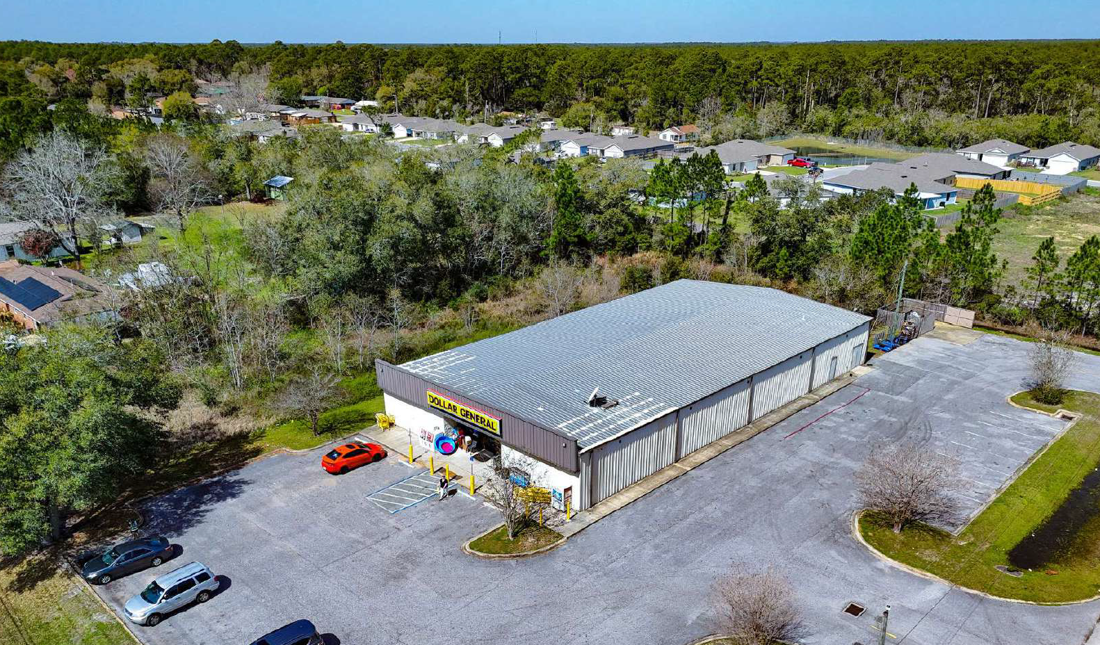 A large single-story retail store named Dollar General in a mostly vacant parking lot, surrounded by trees and nearby residential neighborhood, aerial view during daytime.