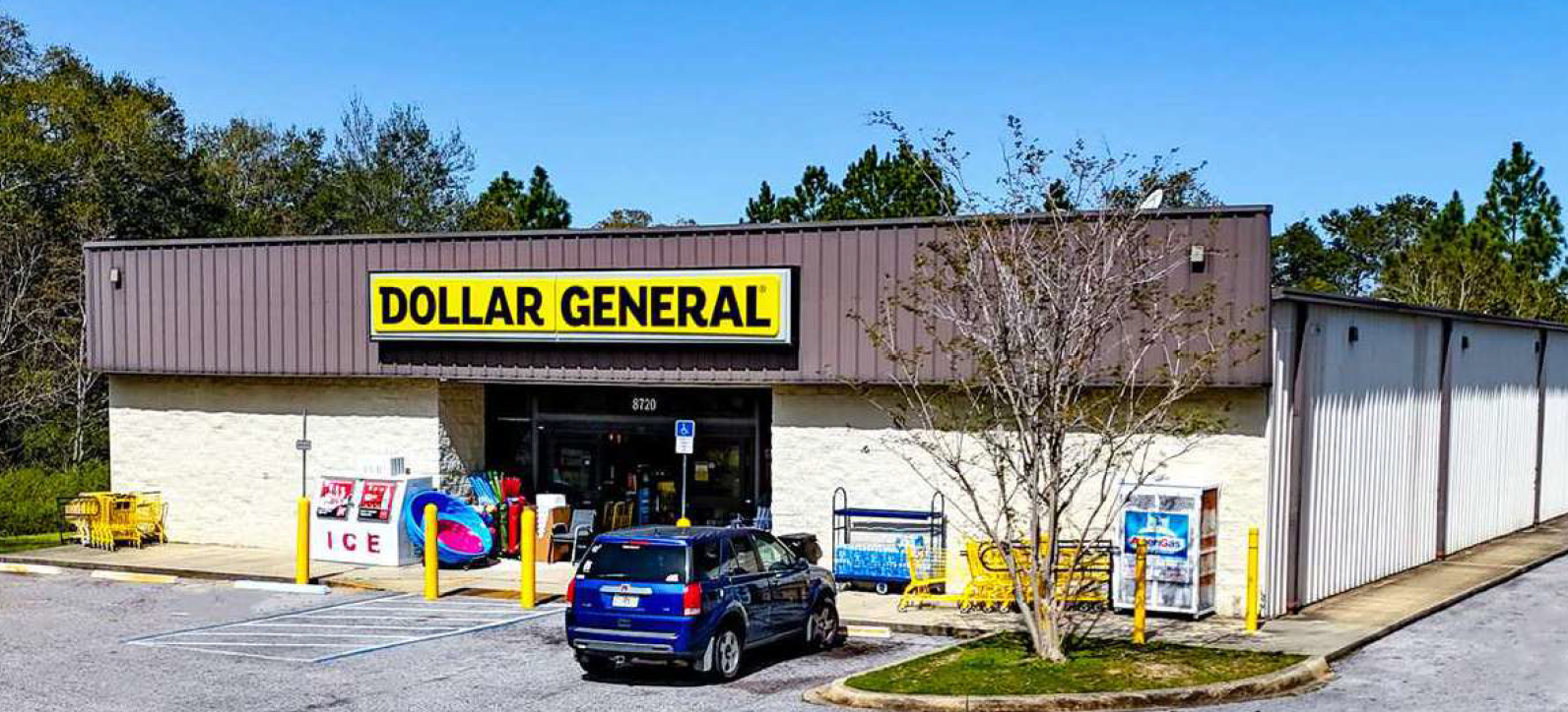 Exterior of a Dollar General store with a parking lot in front, parking spaces, shopping carts, and a blue vehicle parked near the entrance. There is a tree and wooded area behind the store.