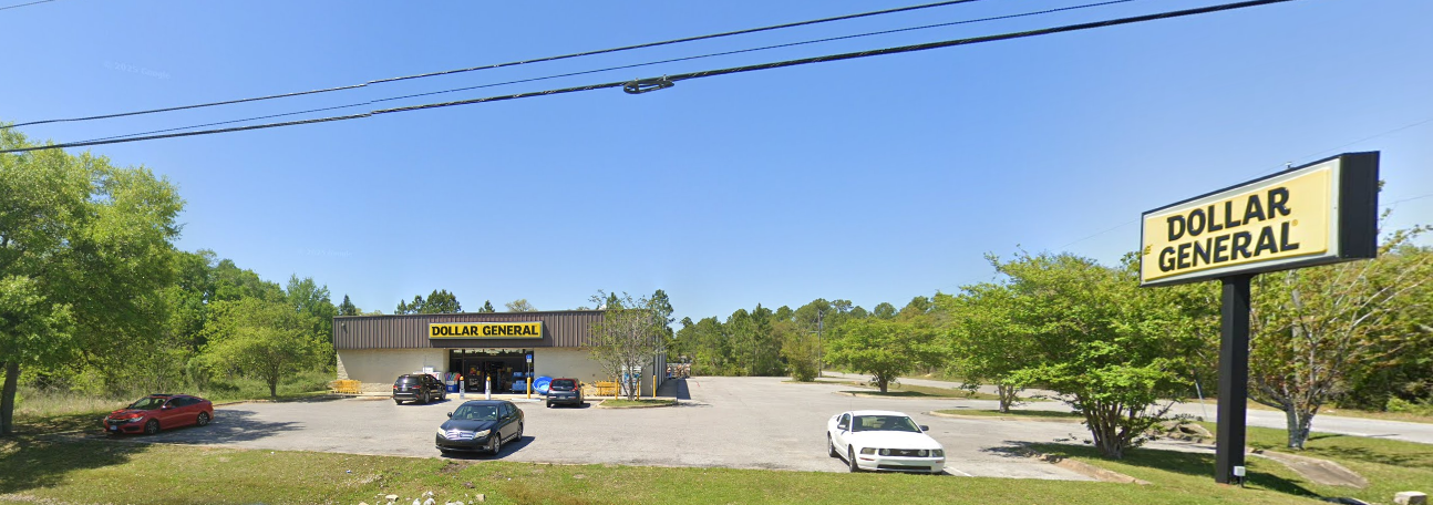A Dollar General store with a parking lot and some cars parked outside, surrounded by green trees under a clear blue sky.