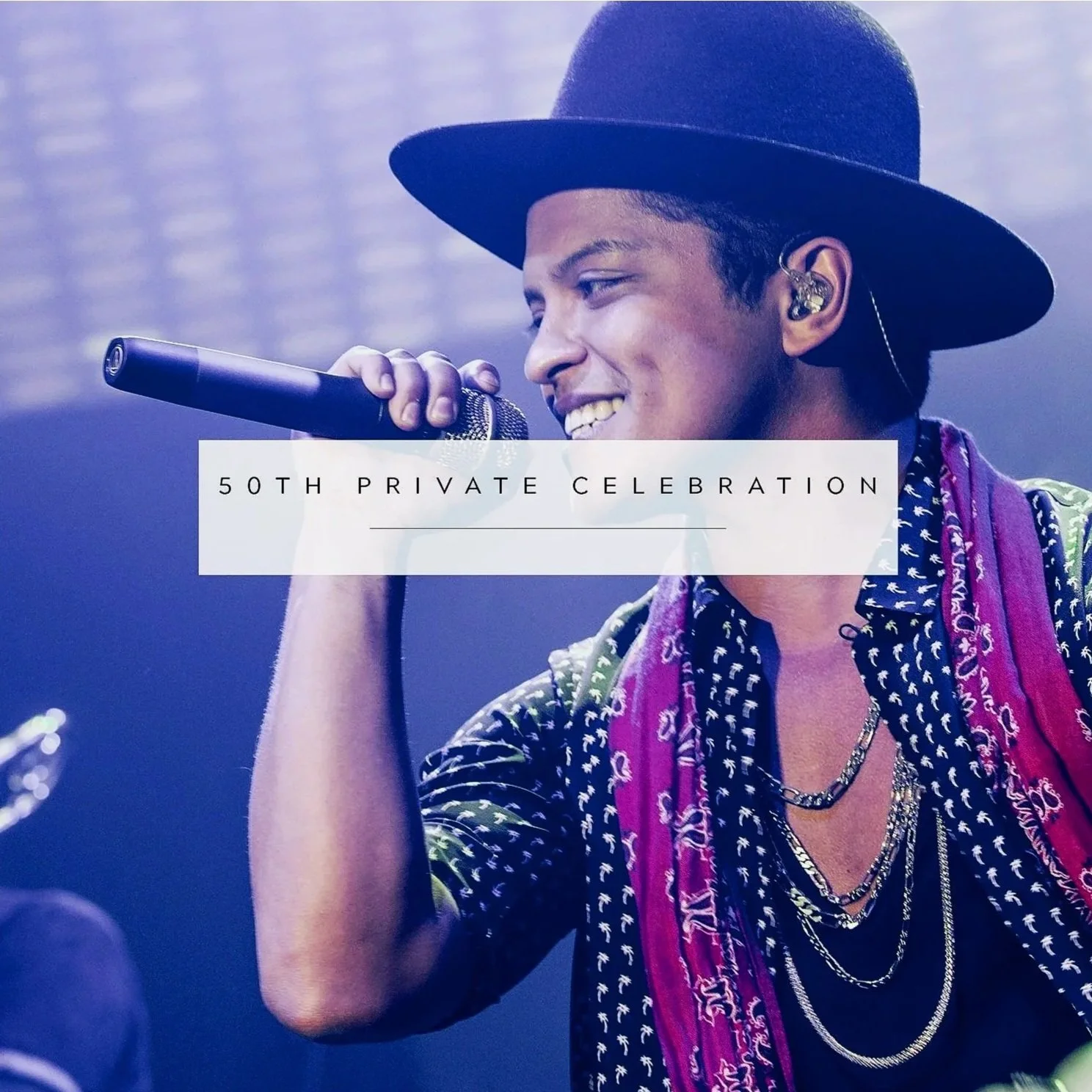 A young male singer wearing a black hat, patterned shirt, and layered necklaces while singing into a microphone at a private celebration event.
