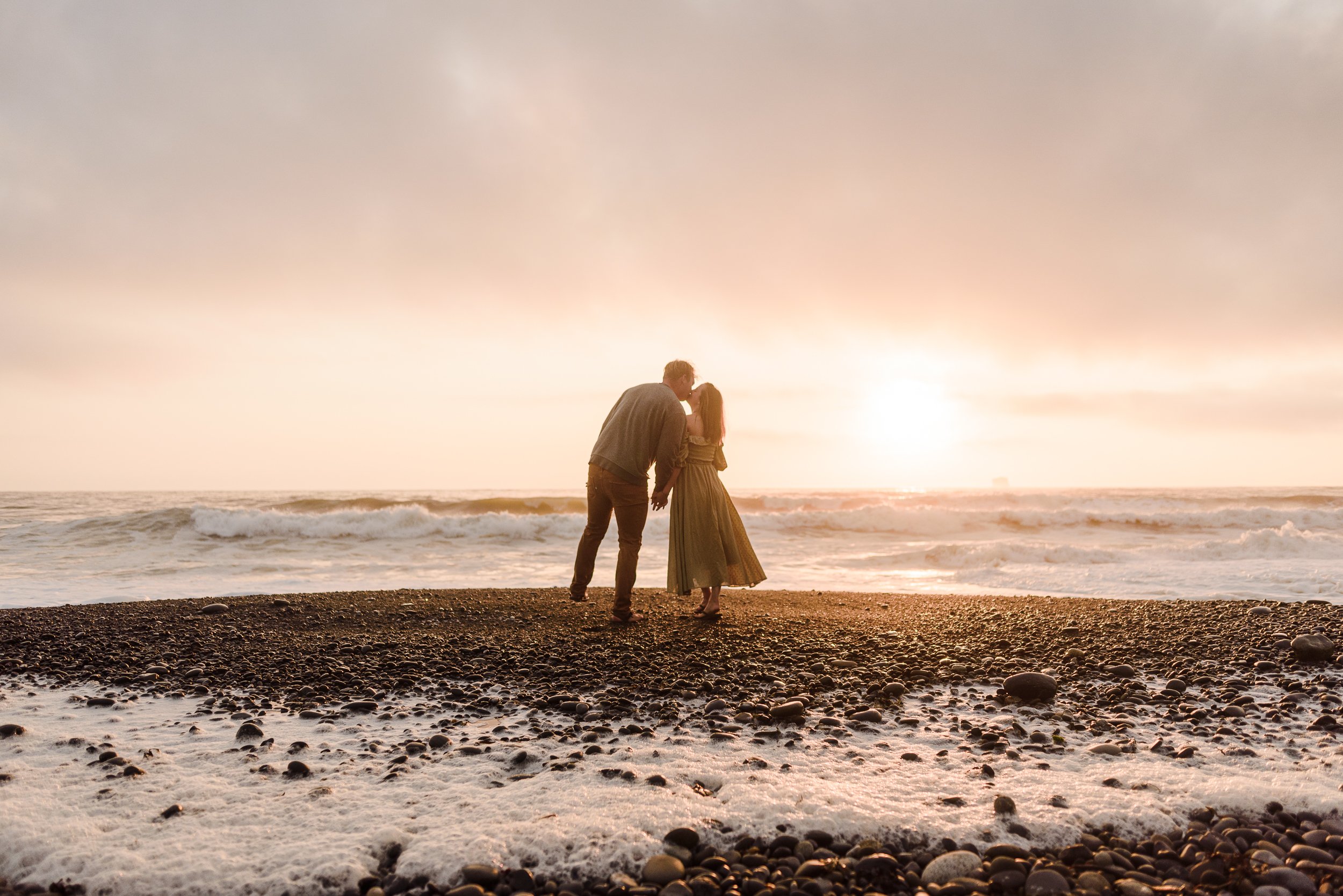 man and woman kissing on rialto beach with ocean in the background
