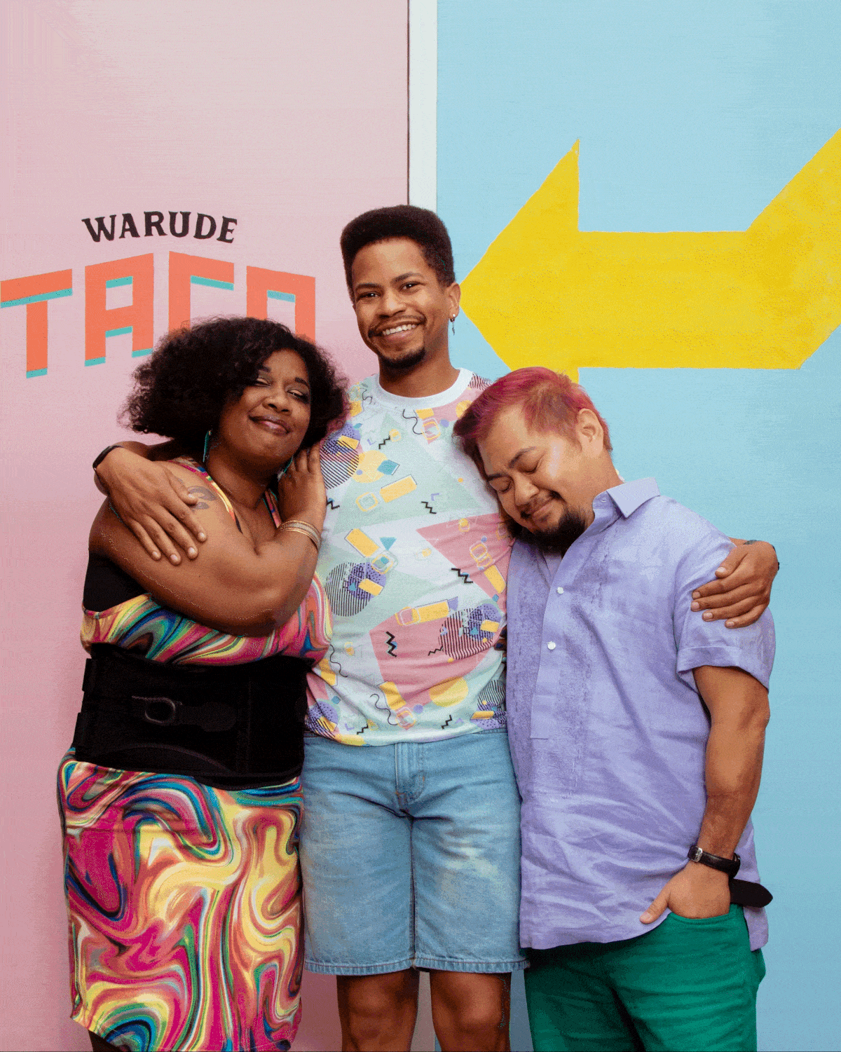 Three people hugging and smiling in front of colorful, geometric background with letters that spell 'WARUDE' and a yellow arrow.
