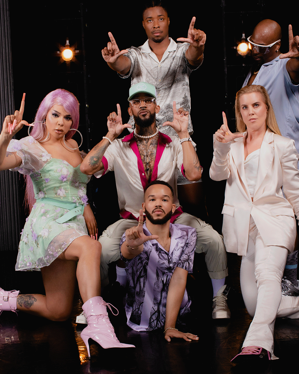 Six diverse people posing with raised hands, making 'L' shapes with their fingers in front of a black background with hanging lights.