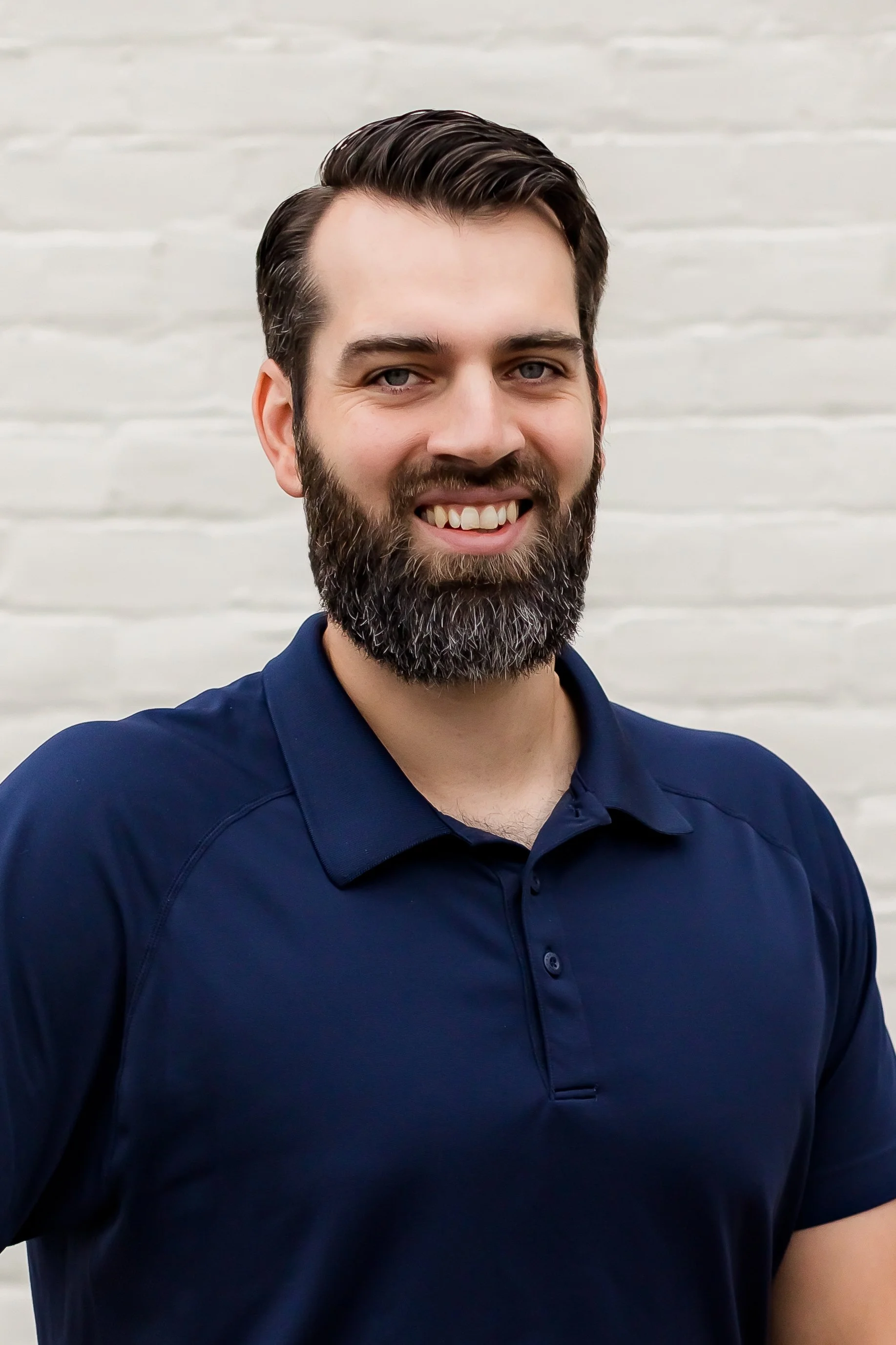 Smiling man with a beard wearing a dark blue polo shirt, standing against a light brick wall background.