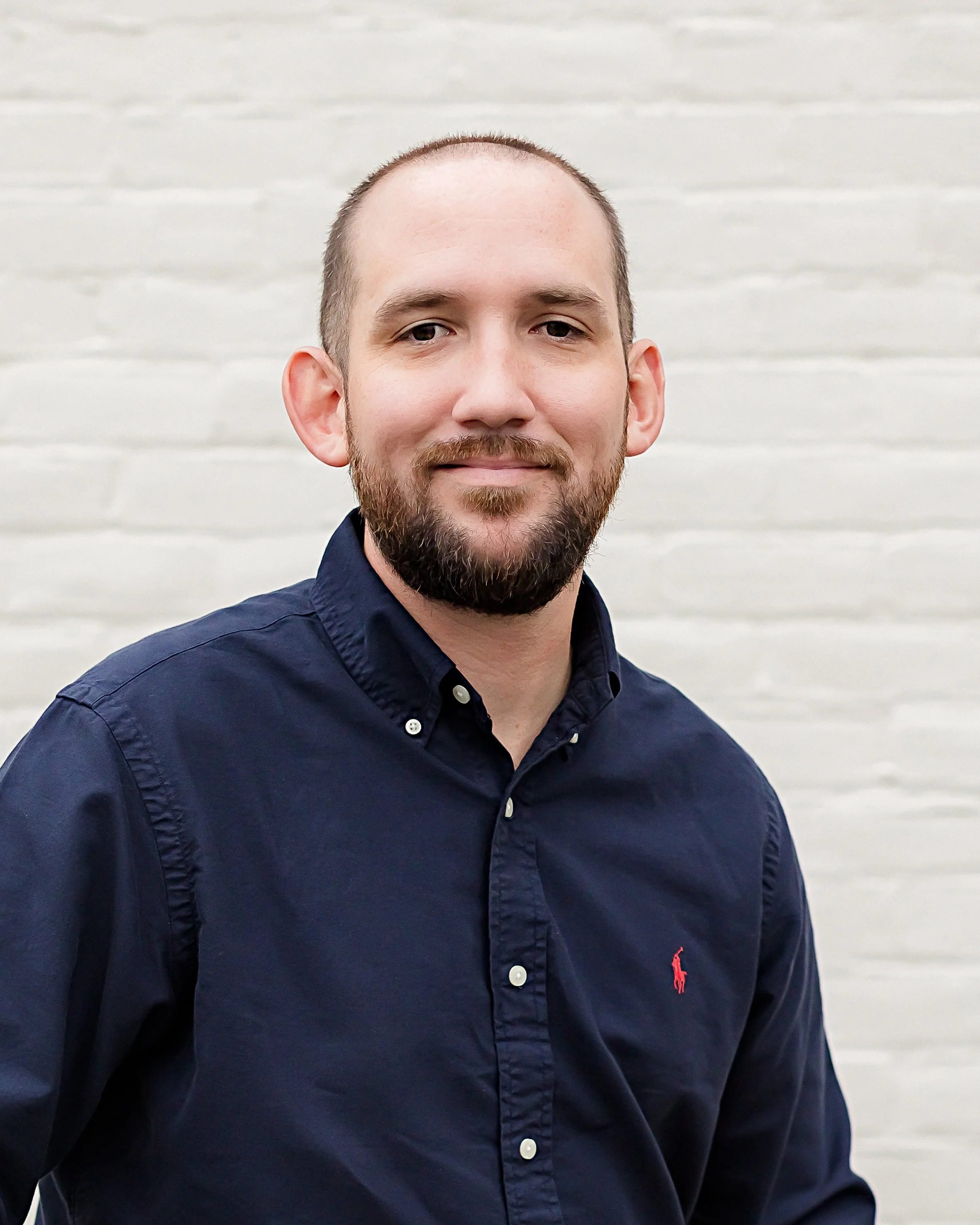 Man wearing a dark navy shirt with a red logo, standing against a light brick wall.