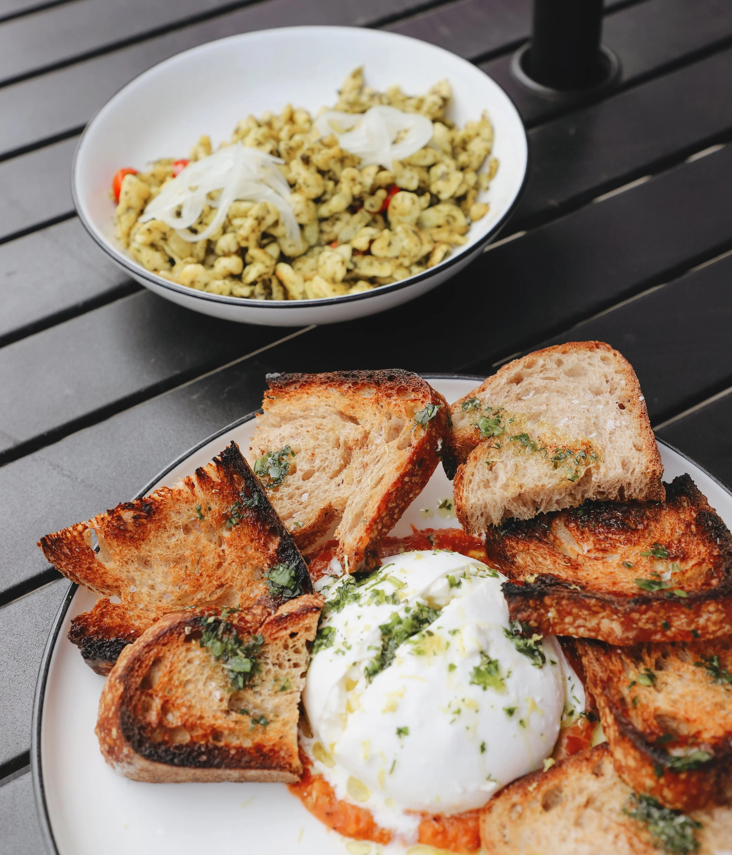 Close-up of a plate with toasted bread slices topped with herbs and a poached egg, next to a bowl of pasta salad with herbs and onions on a black wooden table.