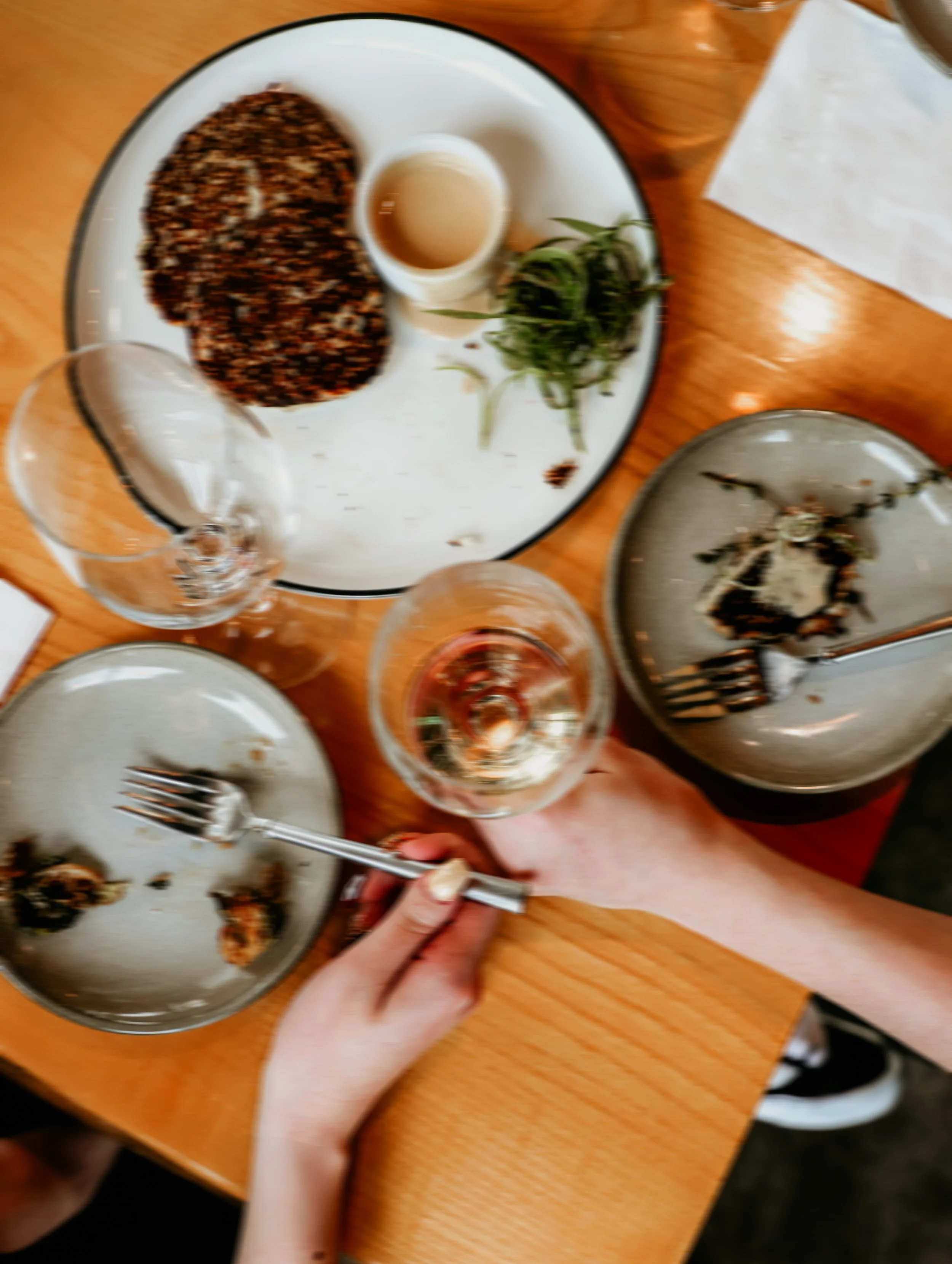 Empty plates, glasses of wine, and a small dish of sauce on a wooden table, with a person holding a fork.