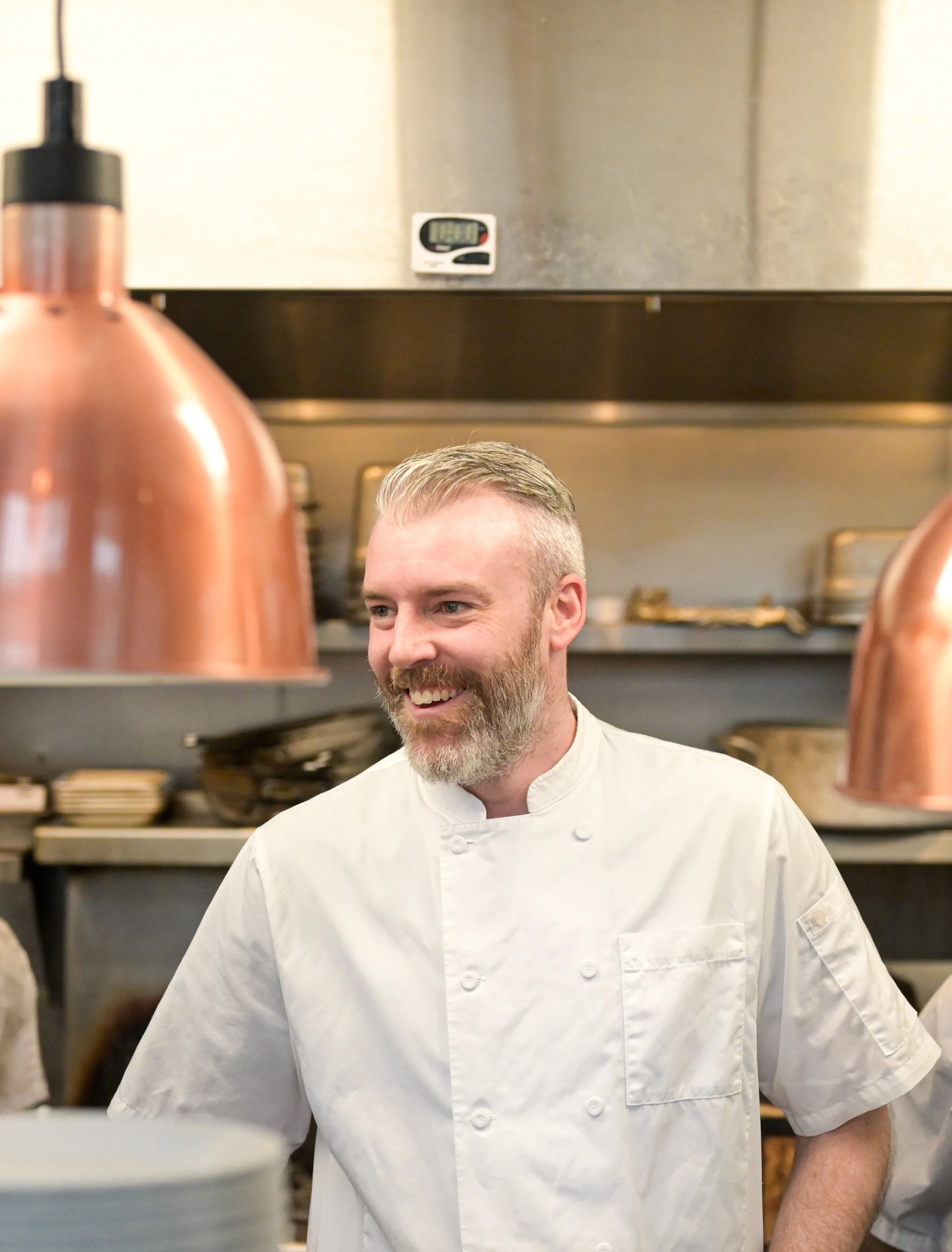 A smiling male chef with a beard wearing a white chef's coat in a professional kitchen.