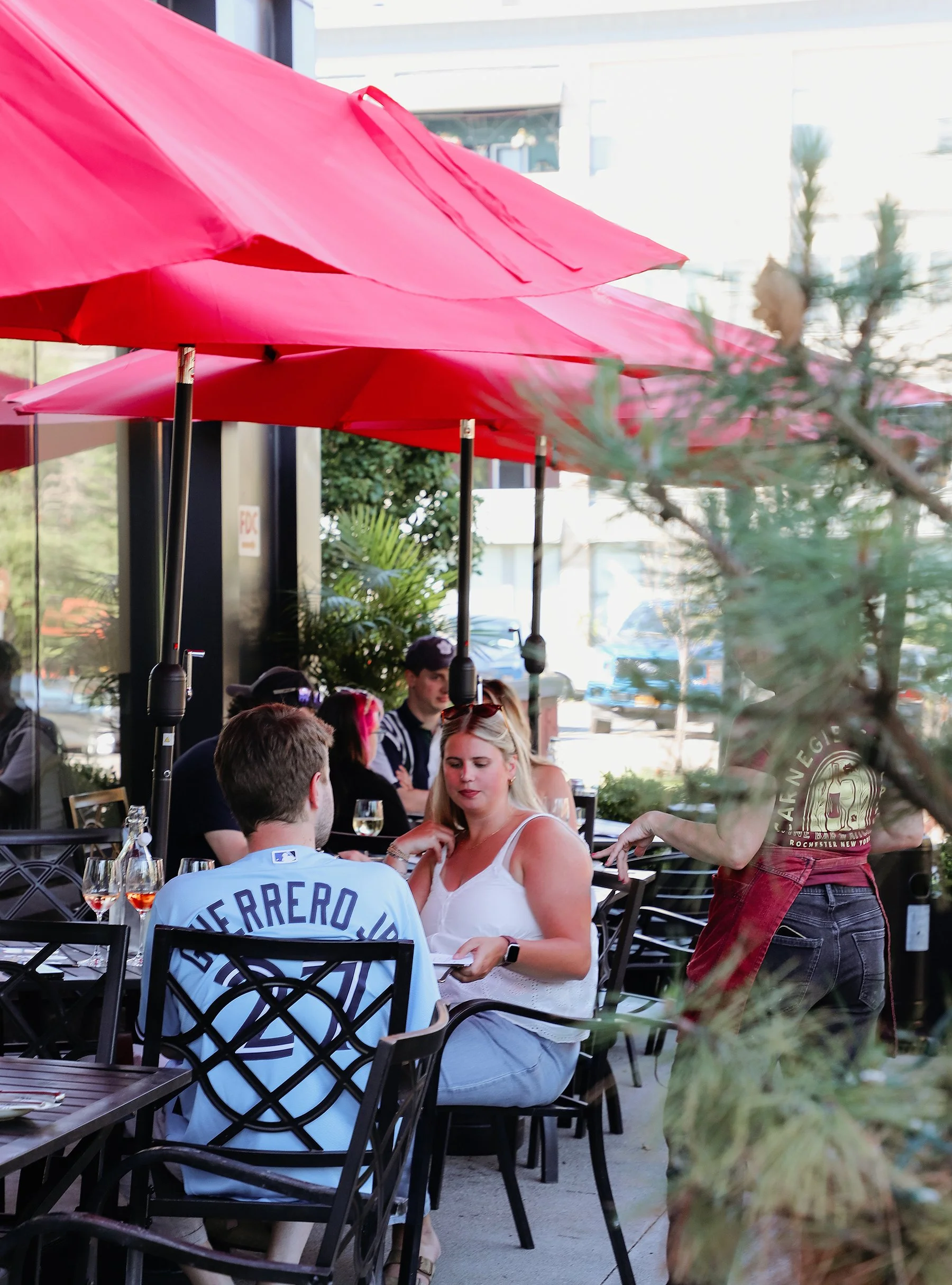 People dining at an outdoor restaurant with red umbrellas, trees, and a city street in the background.
