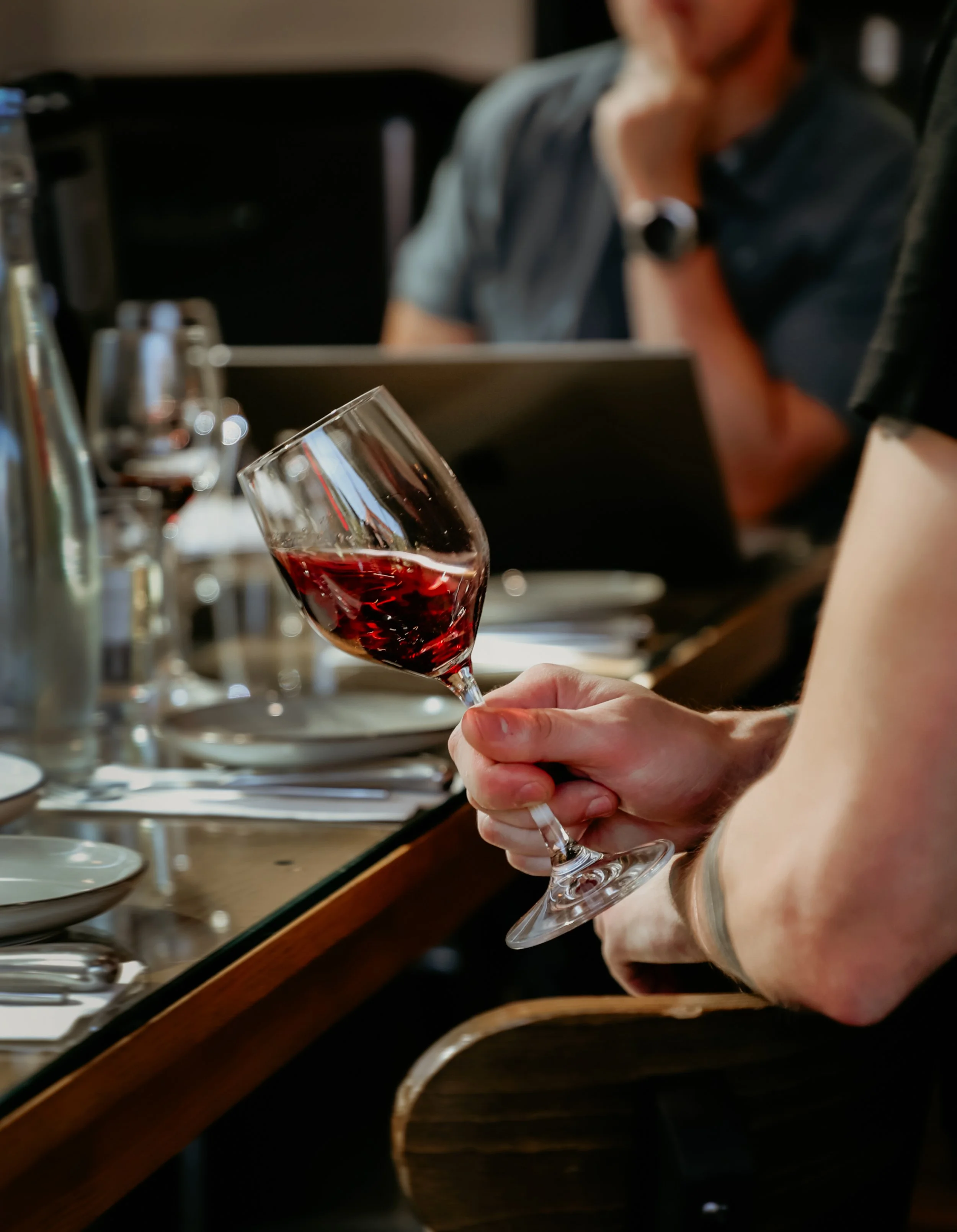 A hand holding a glass of red wine, with a blurred background of a person seated at a table with more wine glasses and bottles.