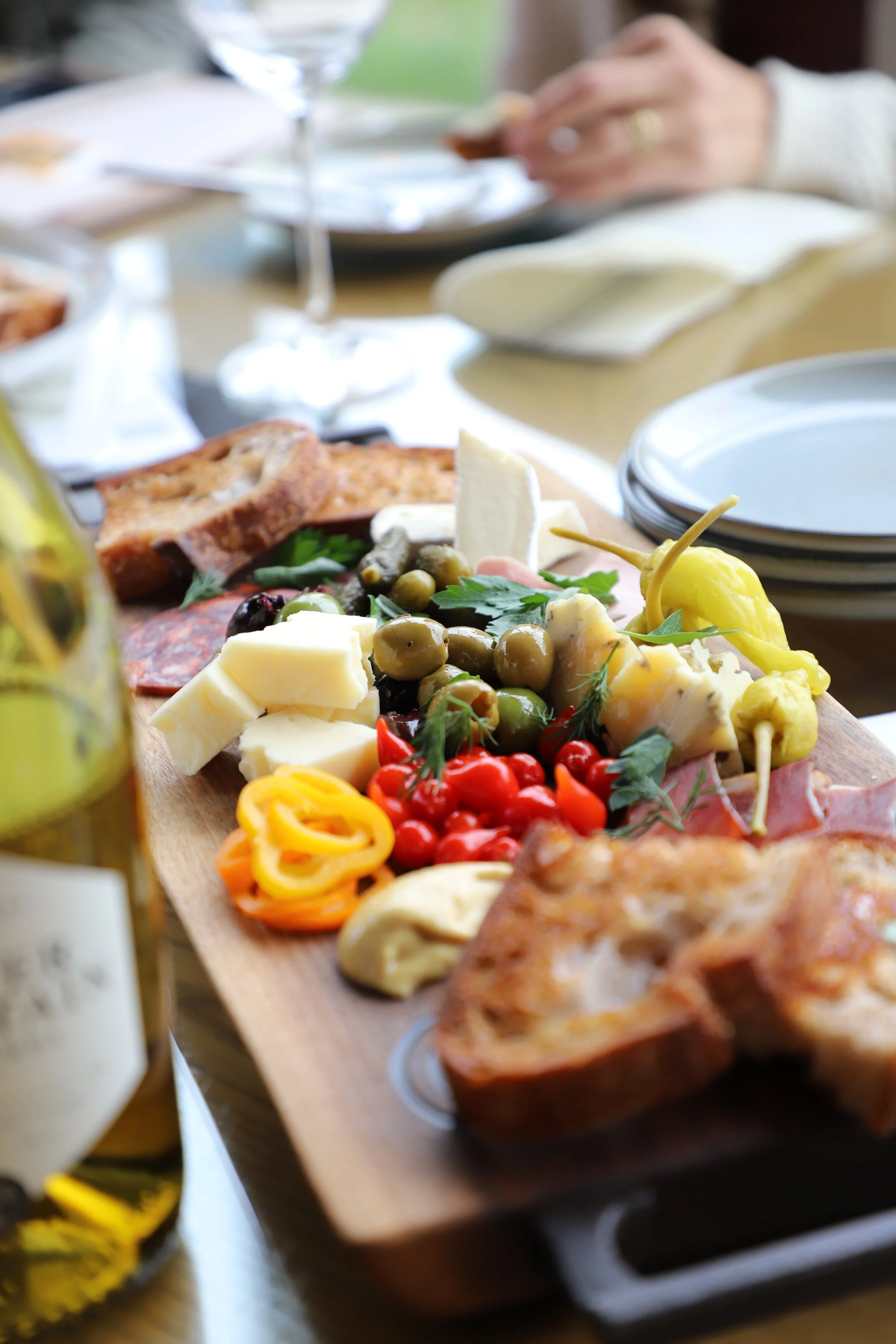 A cheese and charcuterie board with various cheeses, olives, and cured meats on a wooden serving board, with a glass of water, a glass bottle of olive oil, and plates in the background.