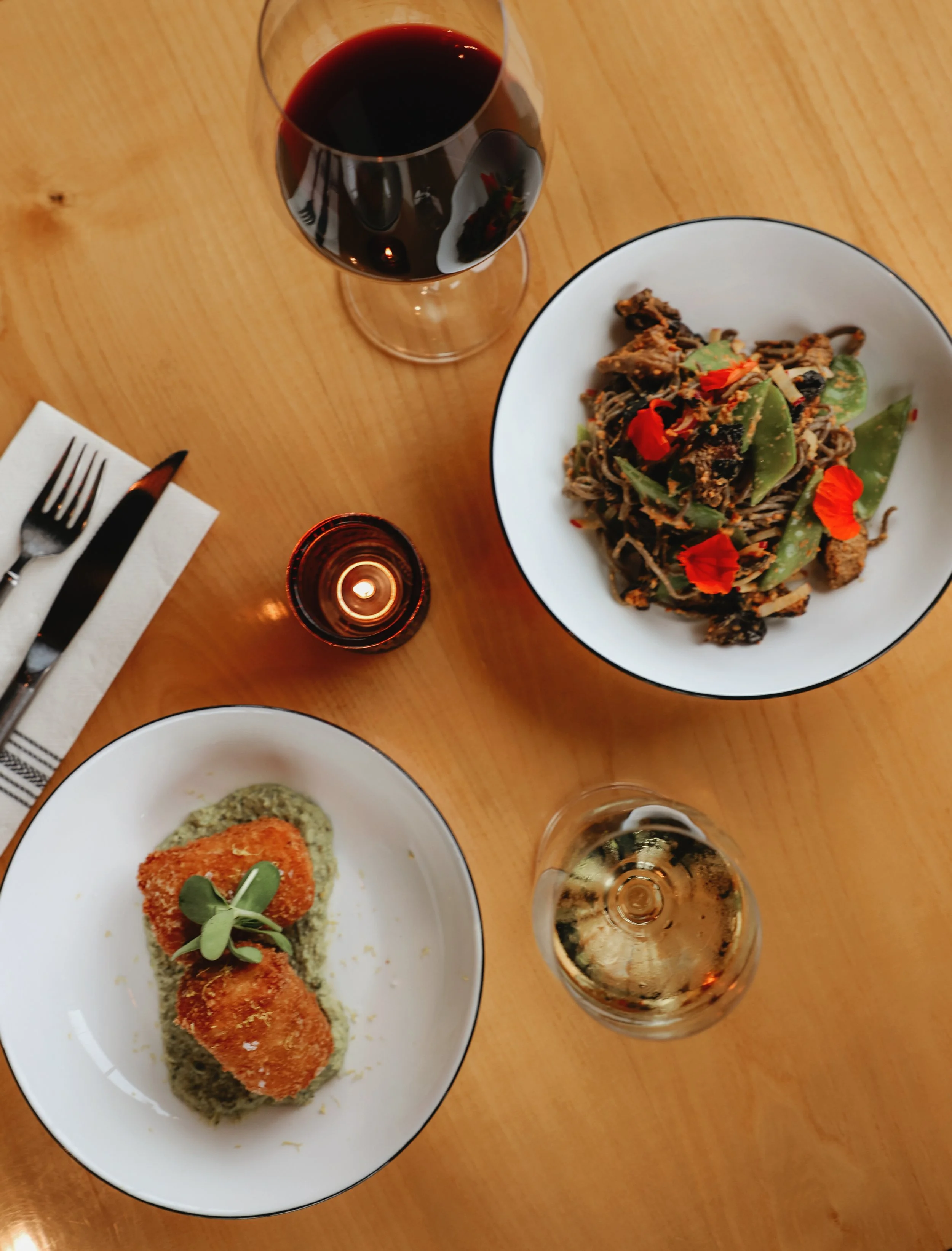 A glass of red wine, a small candle, and two plates of food on a wooden table. One plate has a fried item on a bed of green sauce garnished with microgreens, and the other plate contains a pasta dish with vegetables and edible flowers.