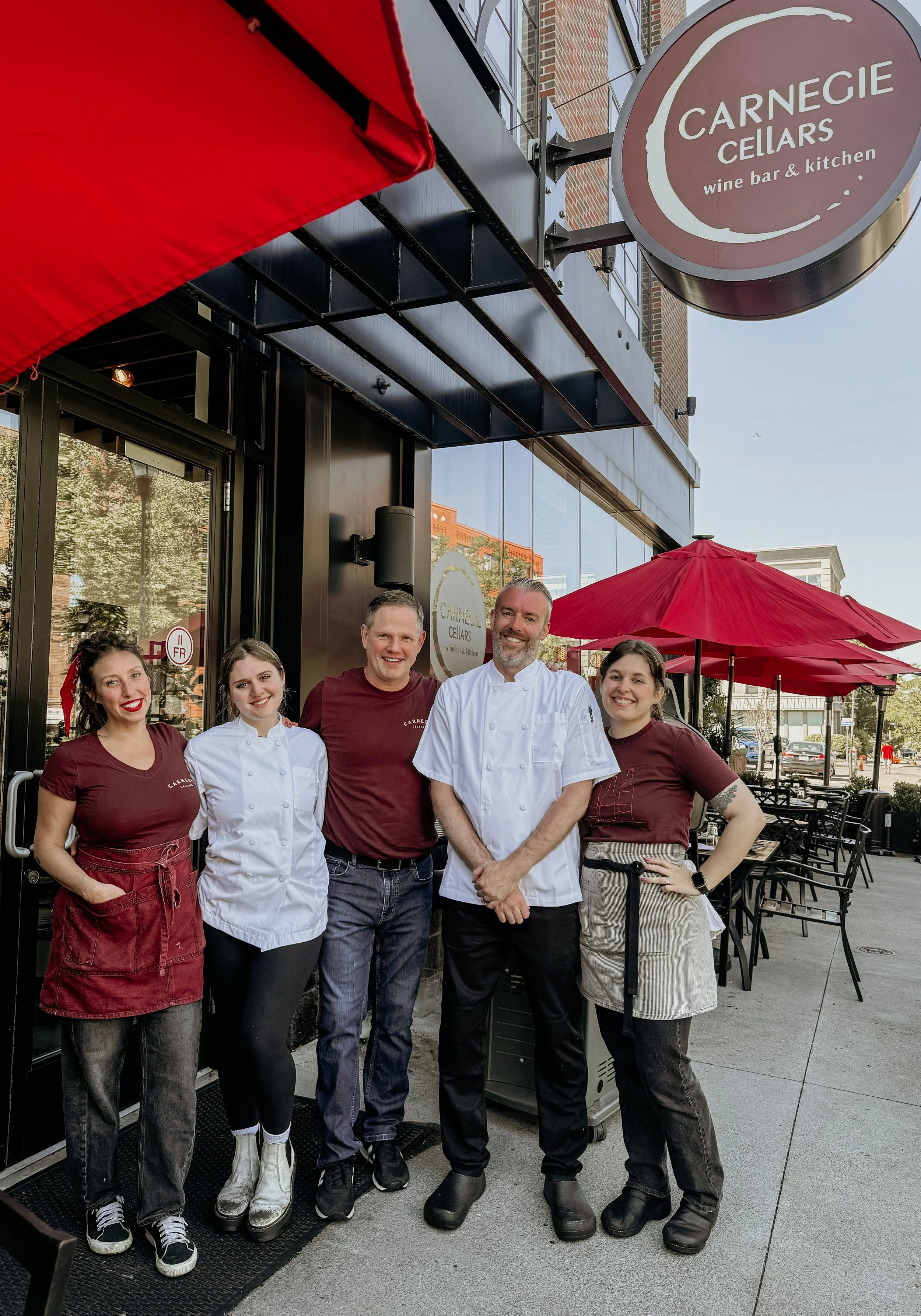 Group of five people standing outside a restaurant called Carnegie Cellars, with red umbrellas and outdoor seating, smiling at the camera.