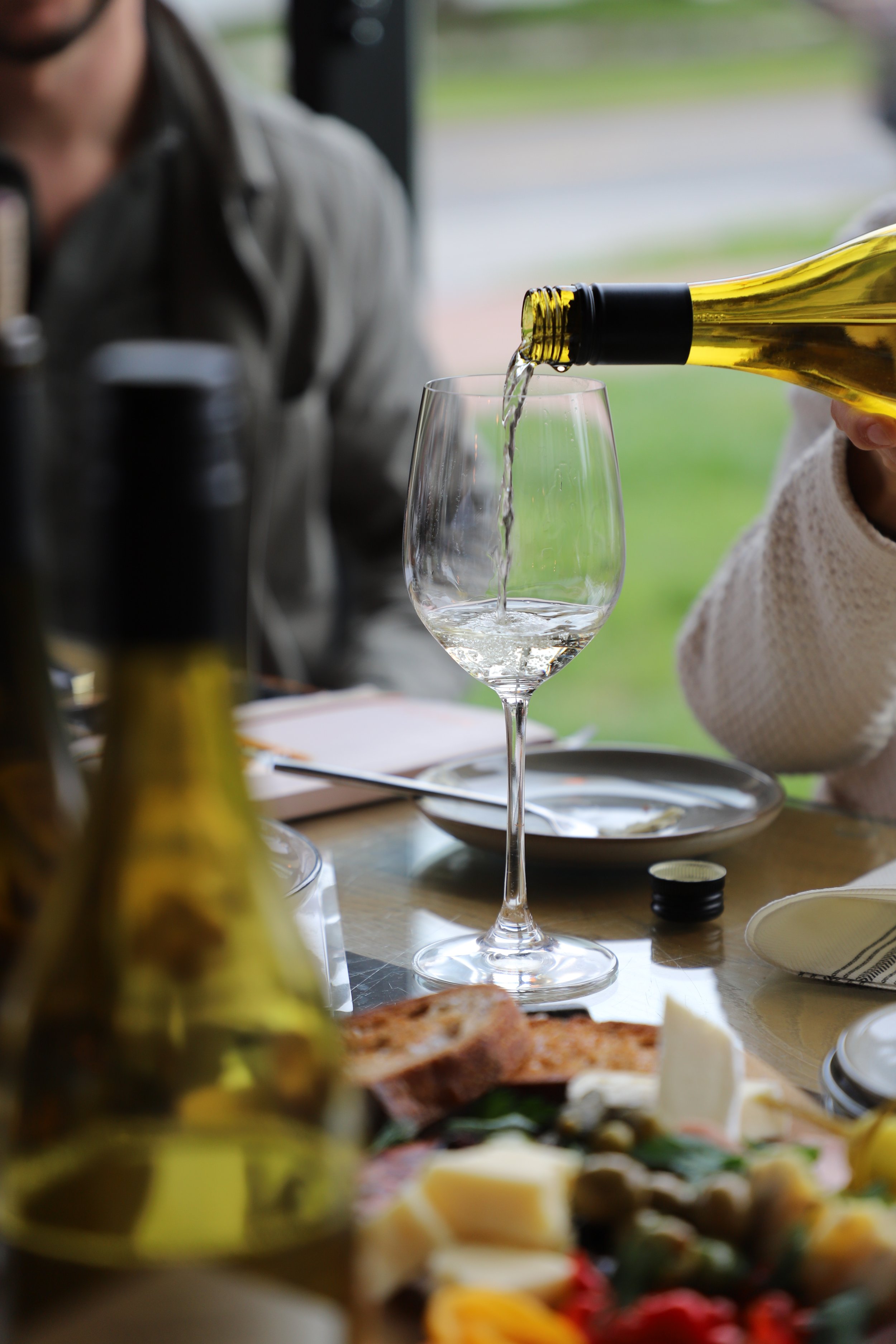 A person pouring white wine into a glass, with a blurred background of a dining table set with food and drink during daytime.