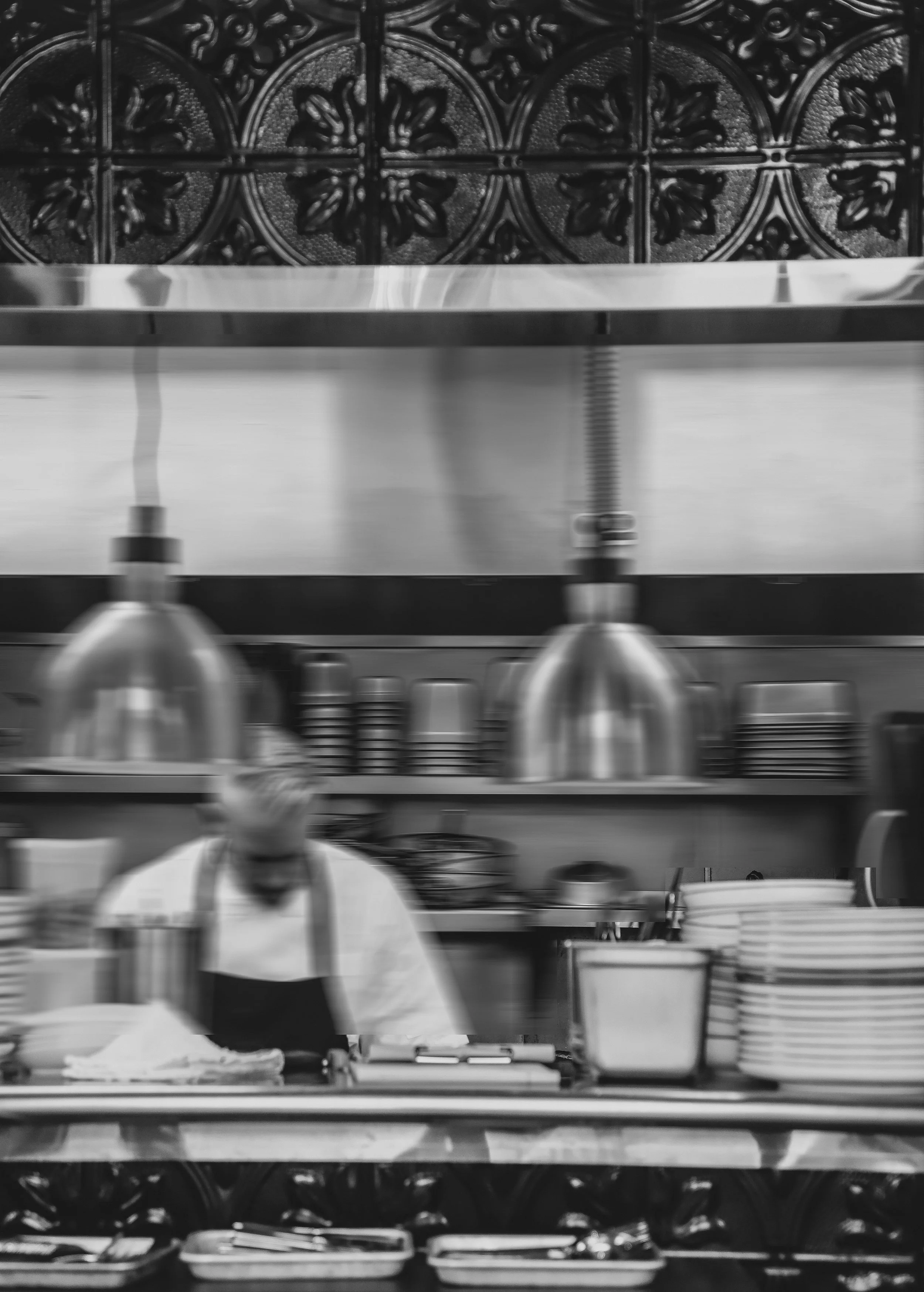 Chef Brian Arliss wearing a white uniform working at a kitchen counter with stainless steel containers, utensils, and dishes, with a decorative tiled wall in the background.