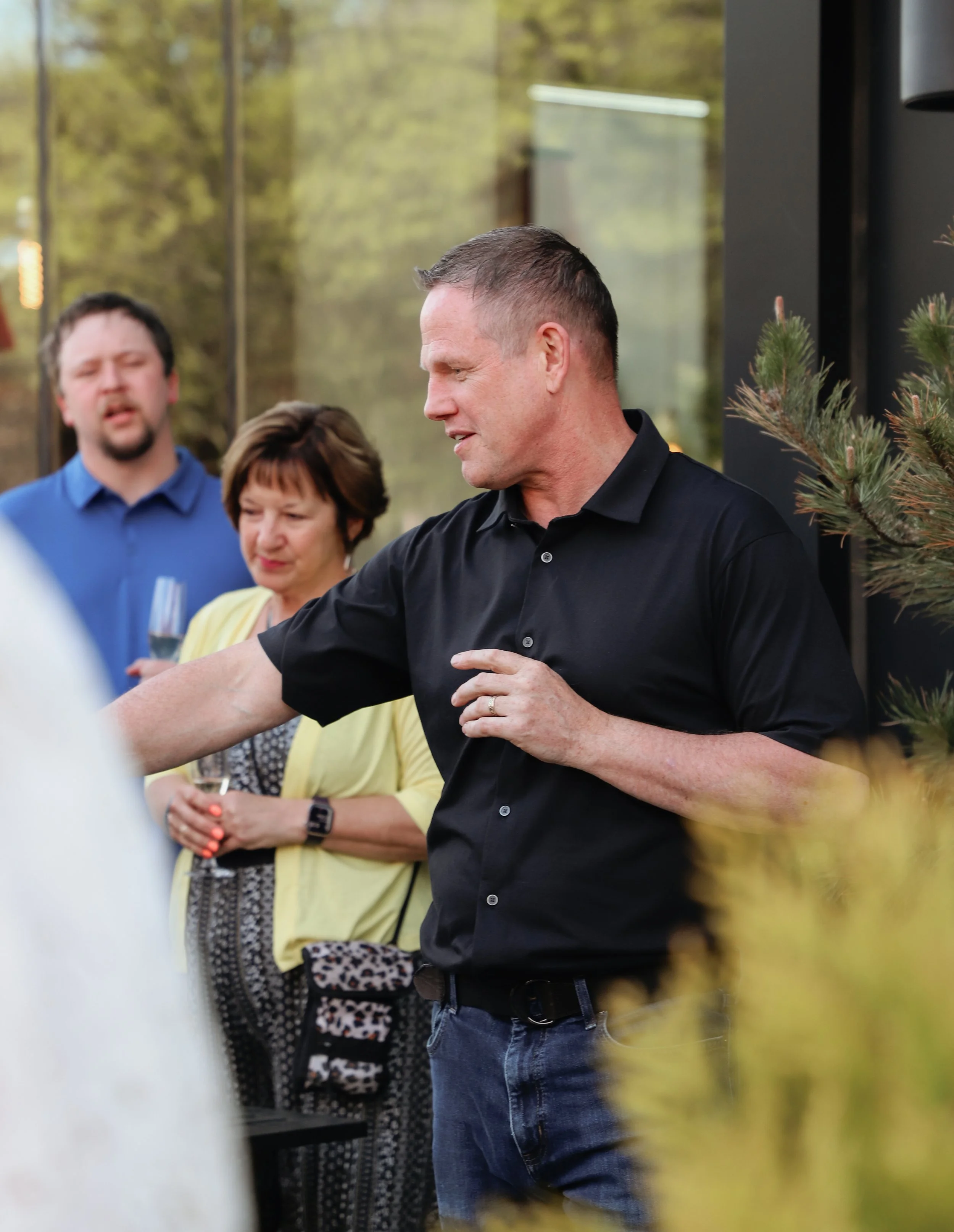Group of people at an outdoor gathering, with one man in a black shirt gesturing and others holding drinks, background of trees and a building.