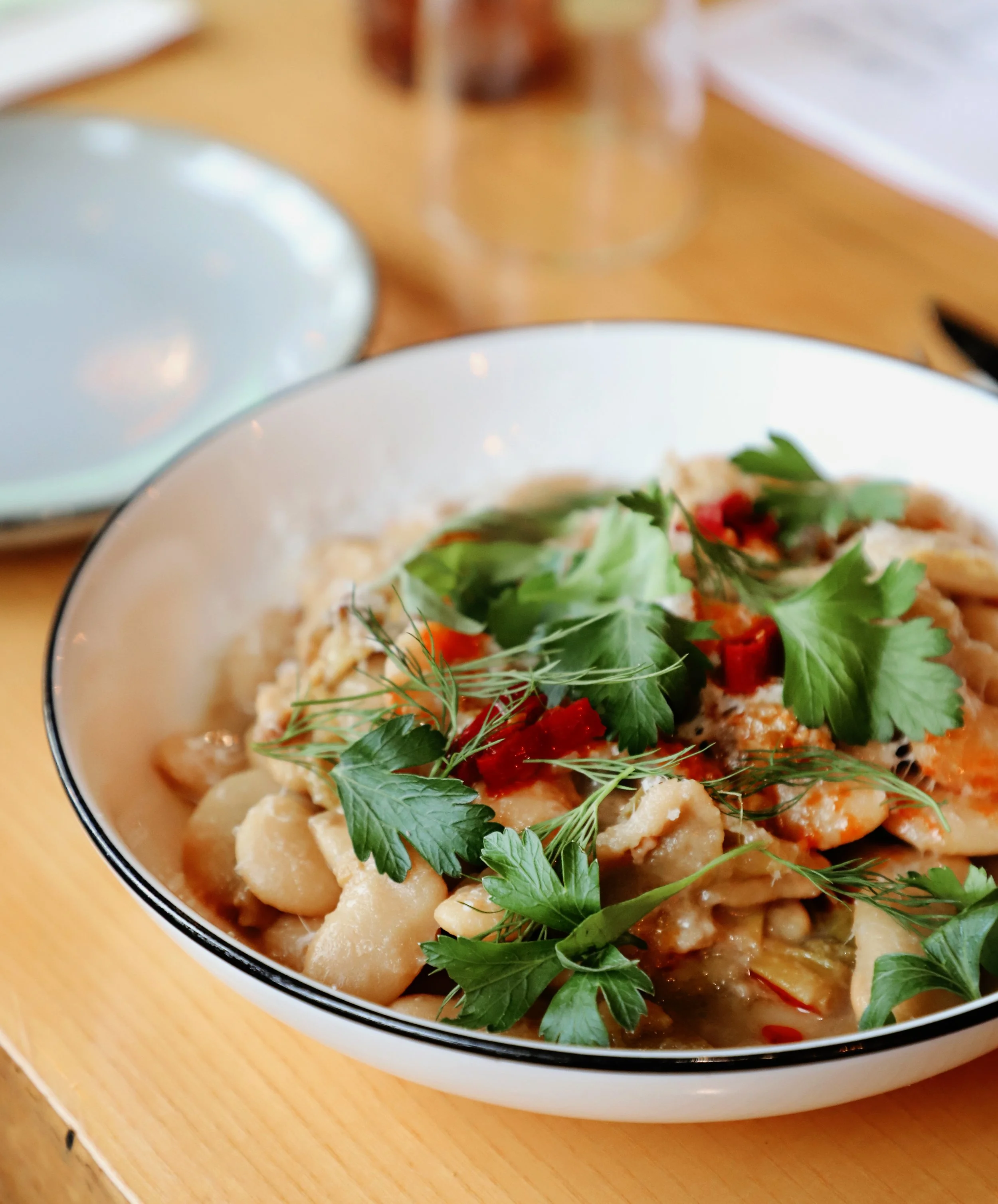 A bowl of food with fresh herbs on top, including cilantro and dill, containing a mix of vegetables and possibly meat or seafood, on a wooden surface with a blurred background.