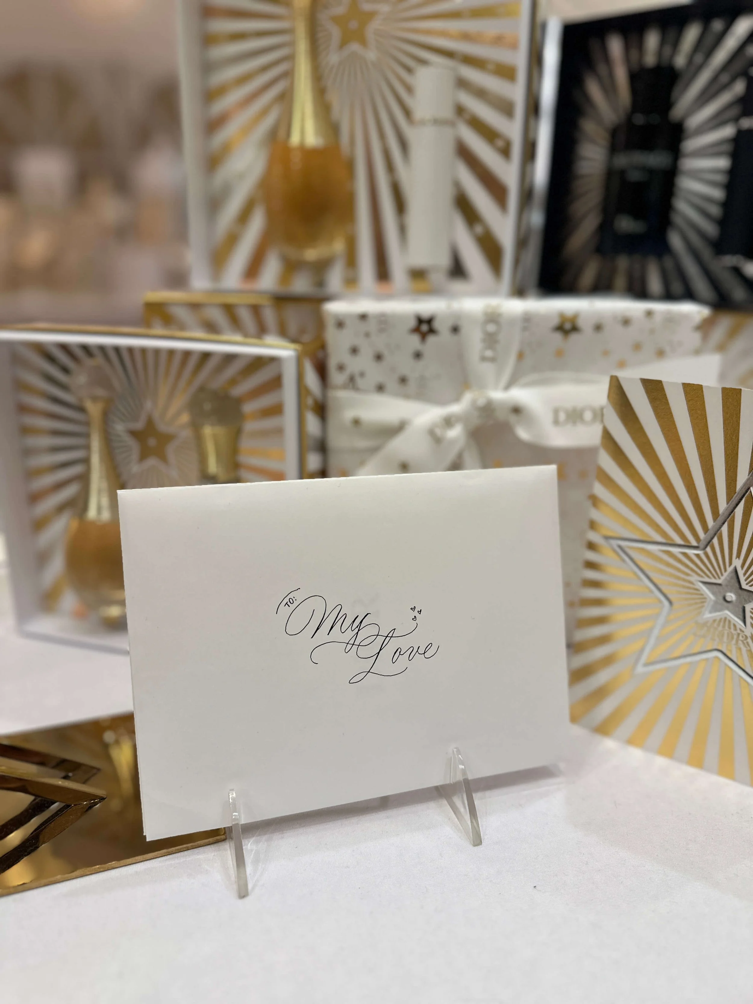 a white envelope with "my love" written in flourished black ink calligraphy on a white table with gold and white Dior perfume boxes obscured in the background