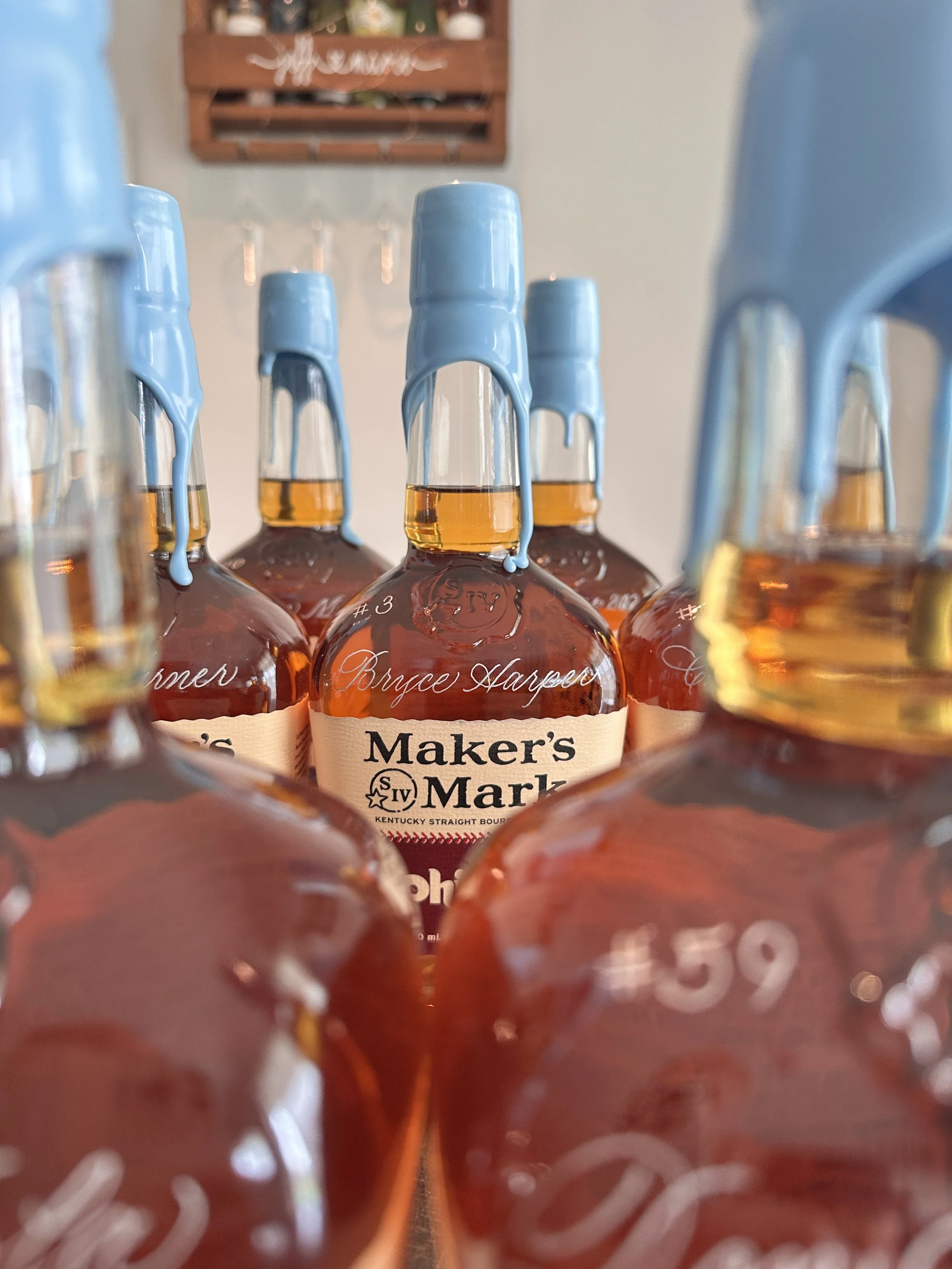 Close-up of several bottles of Maker's Mark bourbon with blue wax seals on the caps, arranged in a row, with a blurred background.
