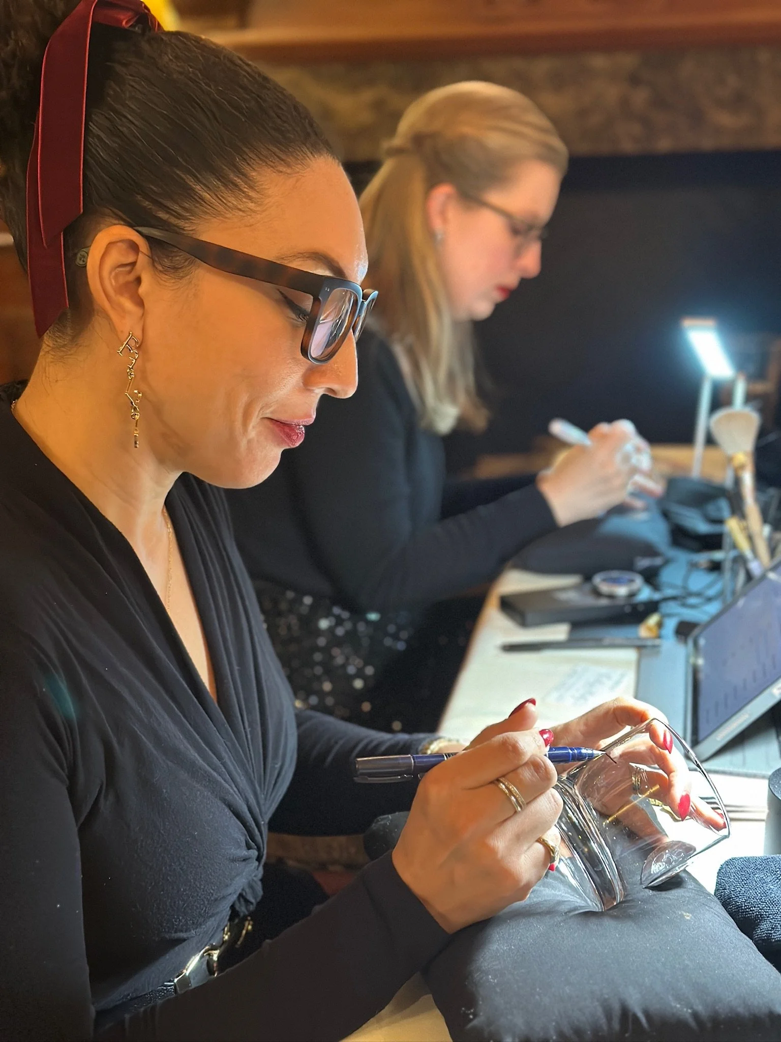 Close-up of a woman wearing glasses and earrings, working on a glass object with a marker, with another woman in the background also focused on her work.