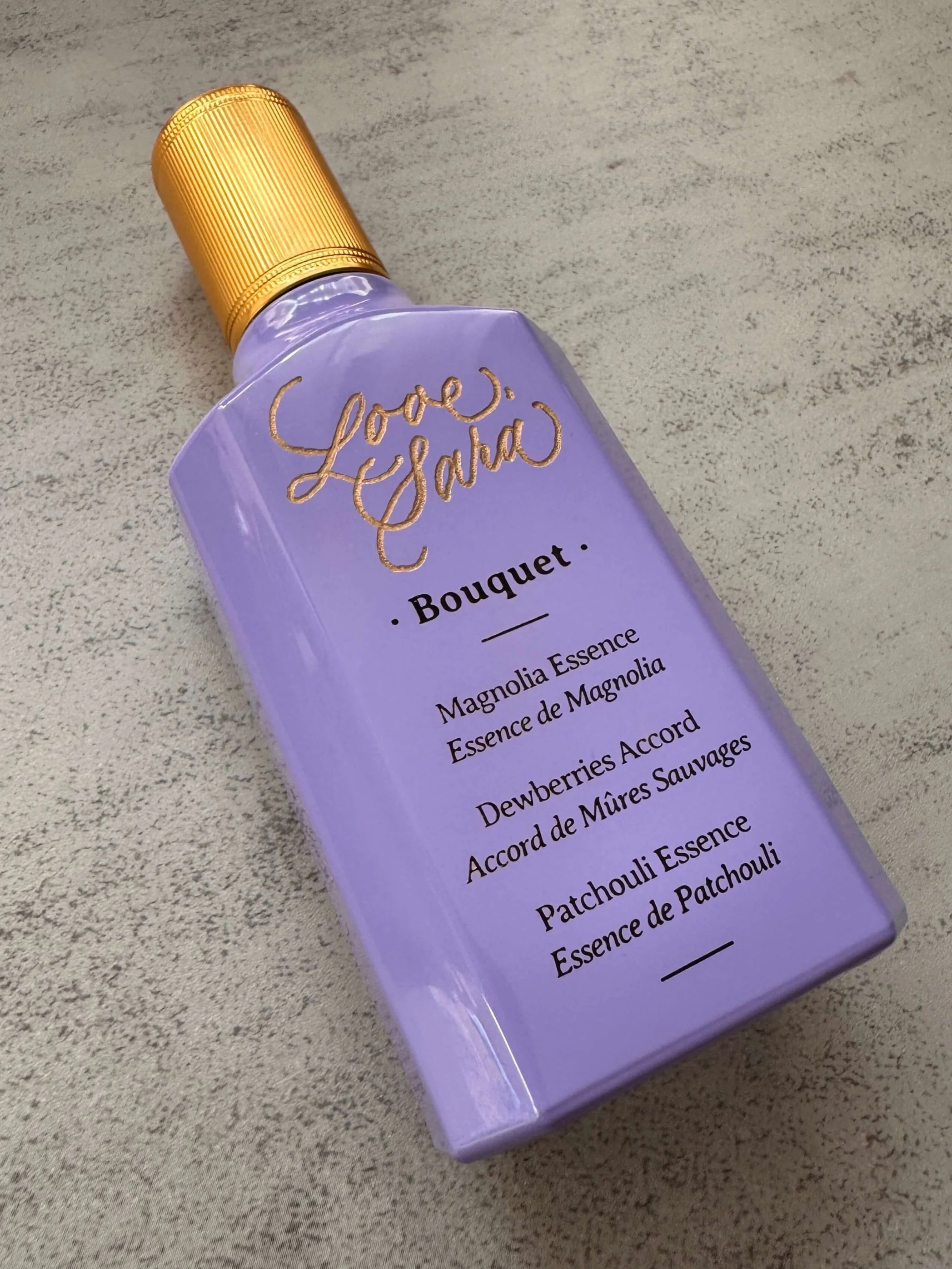 Lavendar perfume bottle with gold lid and gold engraving that reads "Love Sara" on gray background.