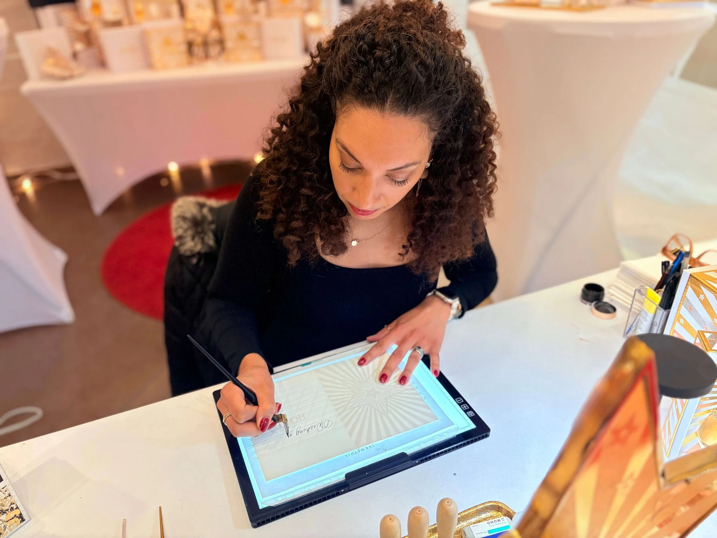 a woman with curly brown hair holding a pointed pen writes in calligraphy on a greeting card upon a white table at which she is seated.
