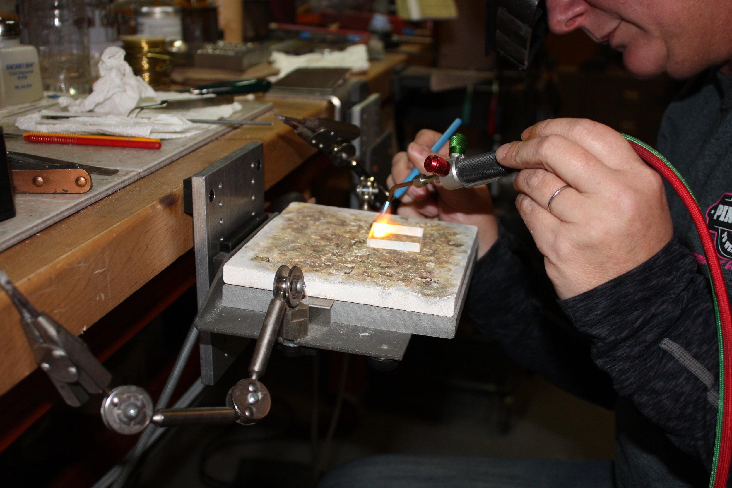 A person working in a workshop with various tools on a cluttered workbench, using a handheld torch or soldering iron.