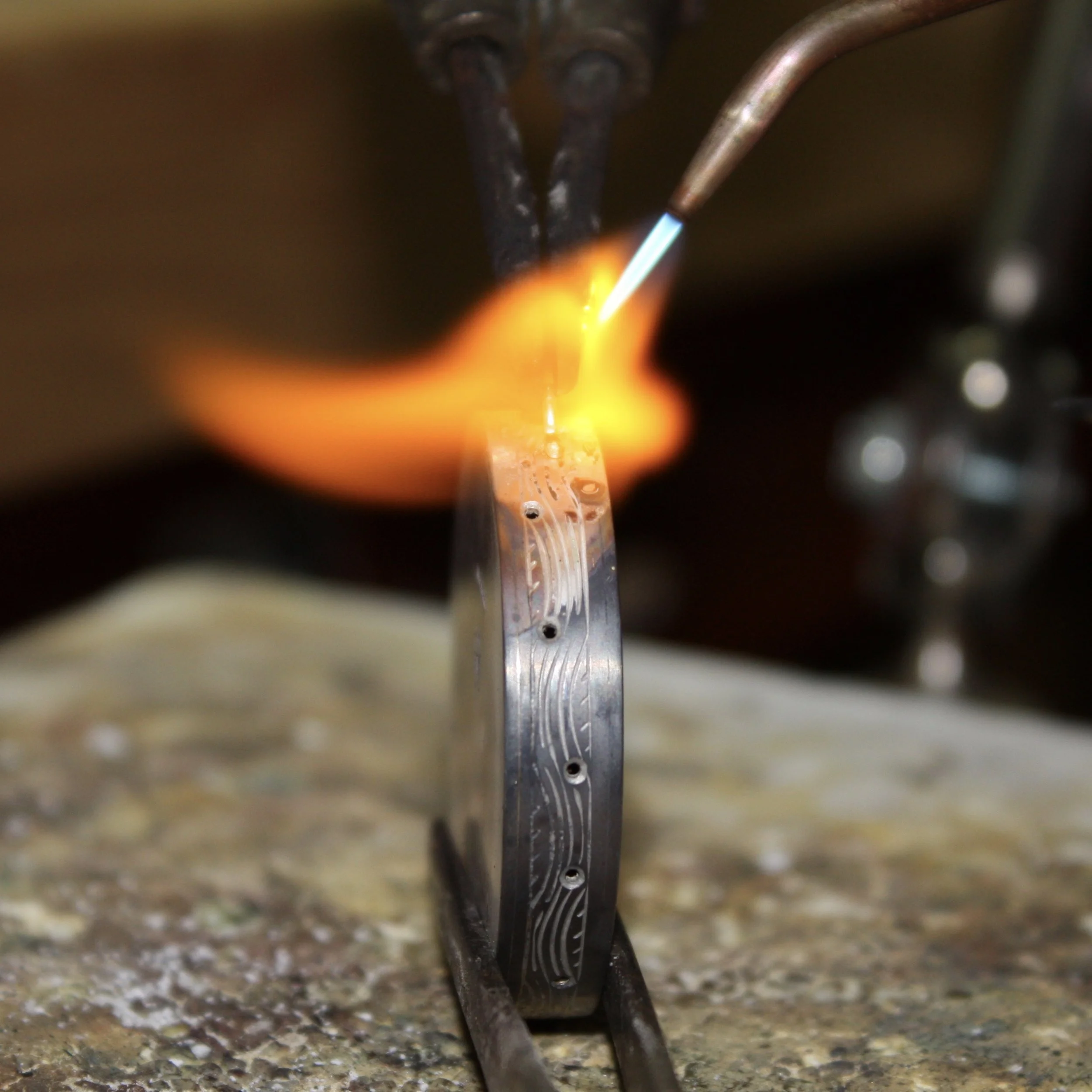 Jeweler heating a textured silver ring with a torch flame at the bench, showing the metal glowing orange during the soldering process.