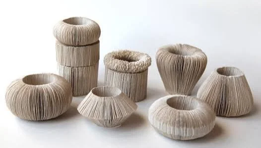 Group of sculptural white and cream fiber vessels with pleated, ribbed surfaces and organic hollow forms arranged against a light background.