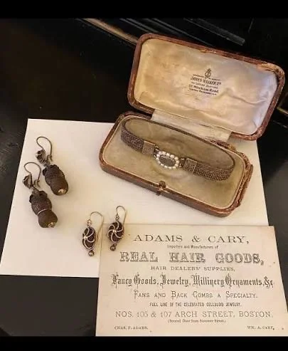 Antique mourning jewelry, including earrings and a bracelet in a fitted case, displayed beside a 19th-century advertisement for hair goods, jewelry, and hairwork supplies.