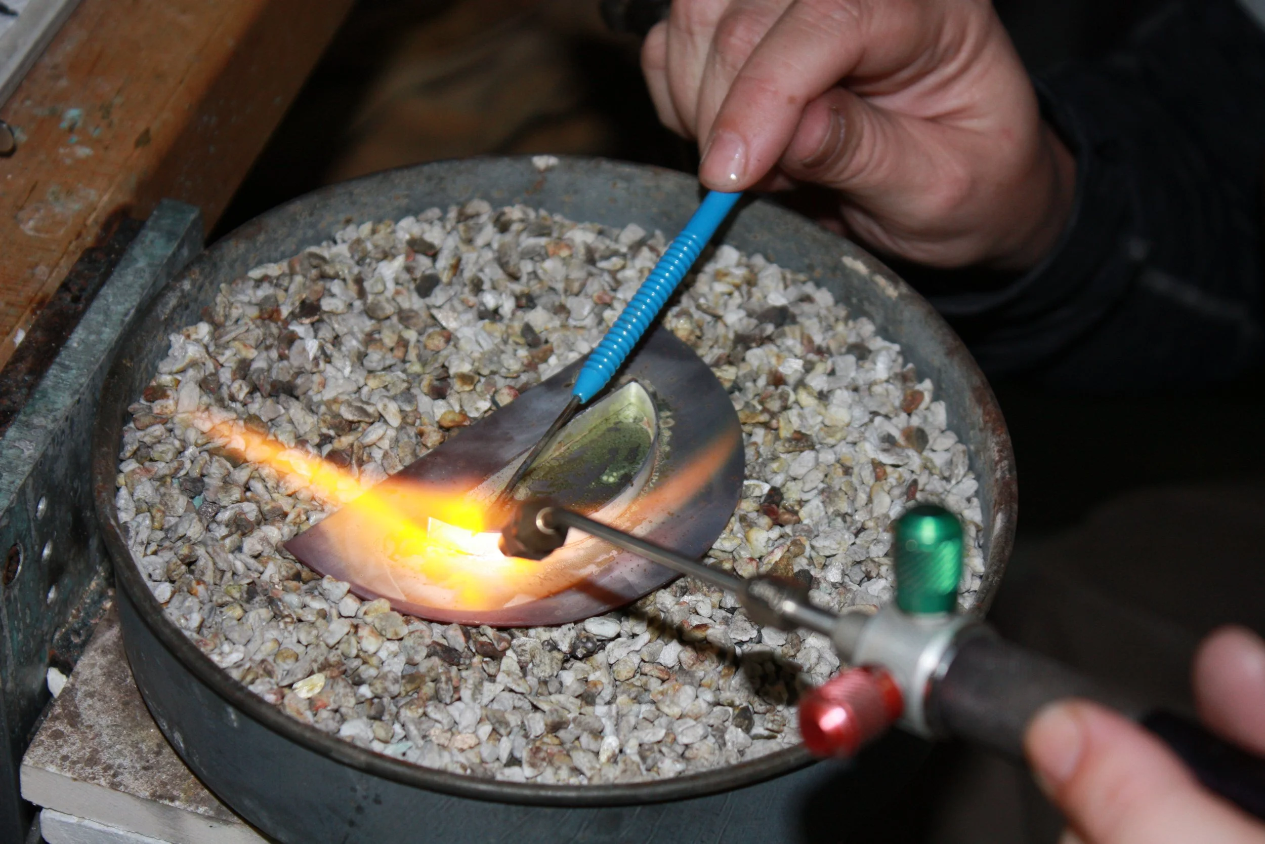 A person using a torch to heat or melt a metal object in a crucible filled with small rocks or gravel. The person is holding the torch in one hand and working over a round, black container on a stove or work surface.