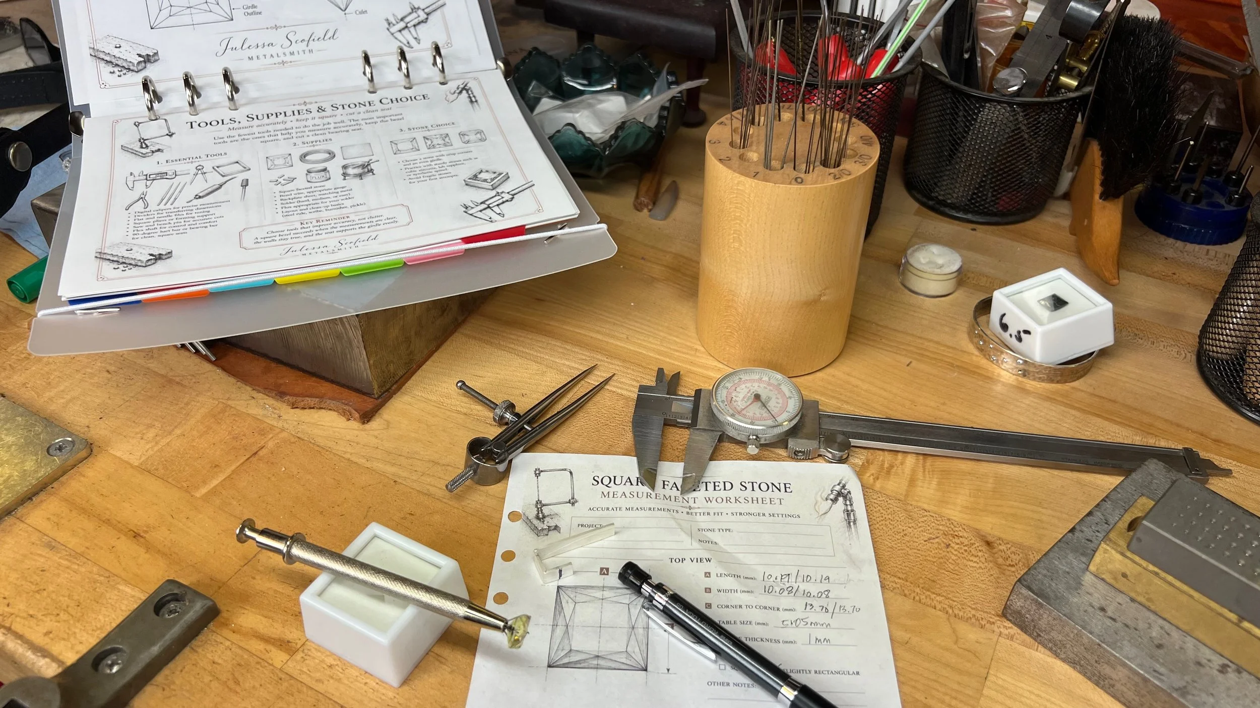 Jewelry tools, dial caliper, square yellow gemstone, and silver bezel material arranged on a metalsmith’s workbench.
