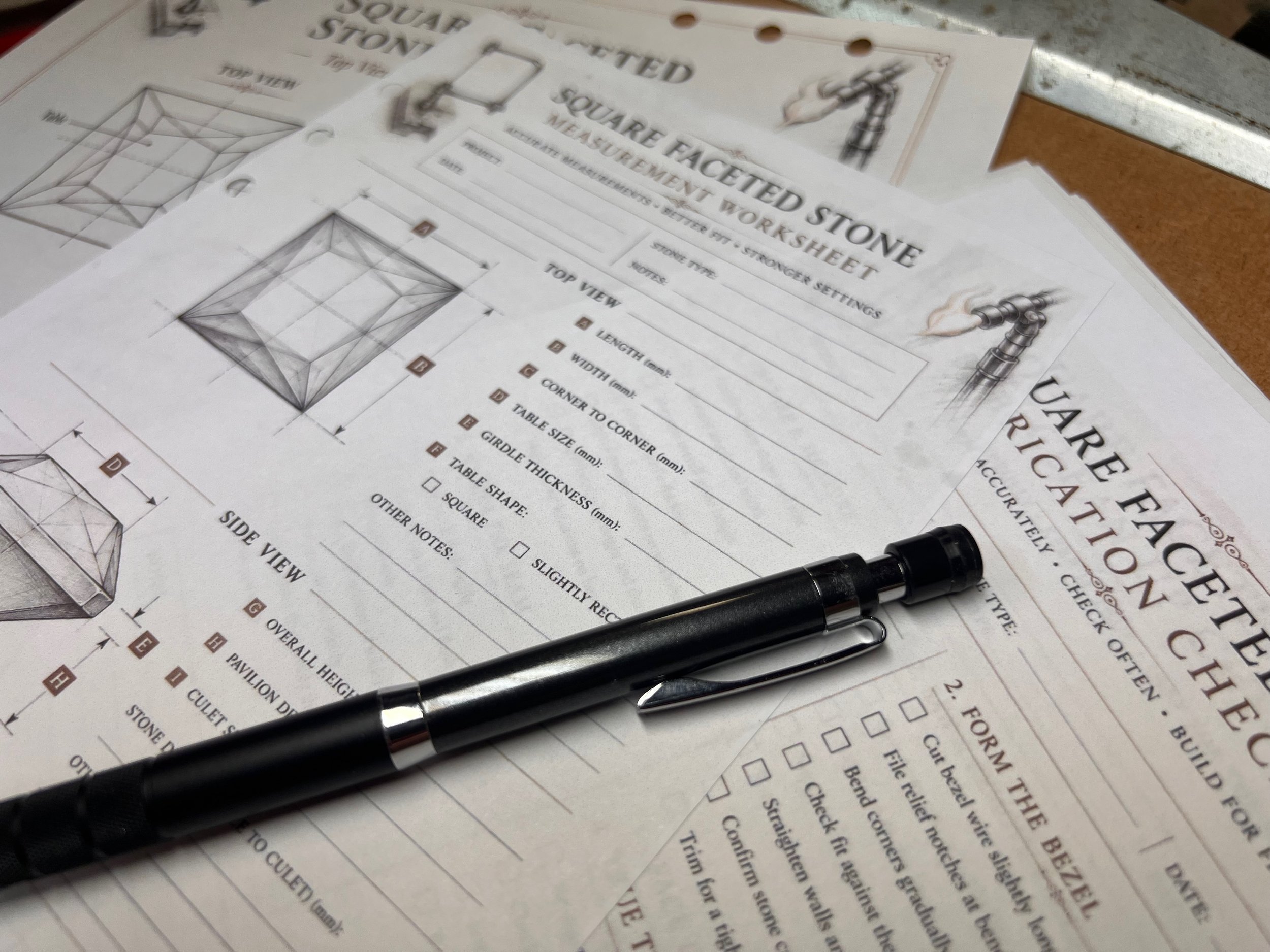 A close-up of engineering worksheets for a square faceted stone, showing diagrams, measurements, and notes, with a black pen and a silver pen resting on the papers.