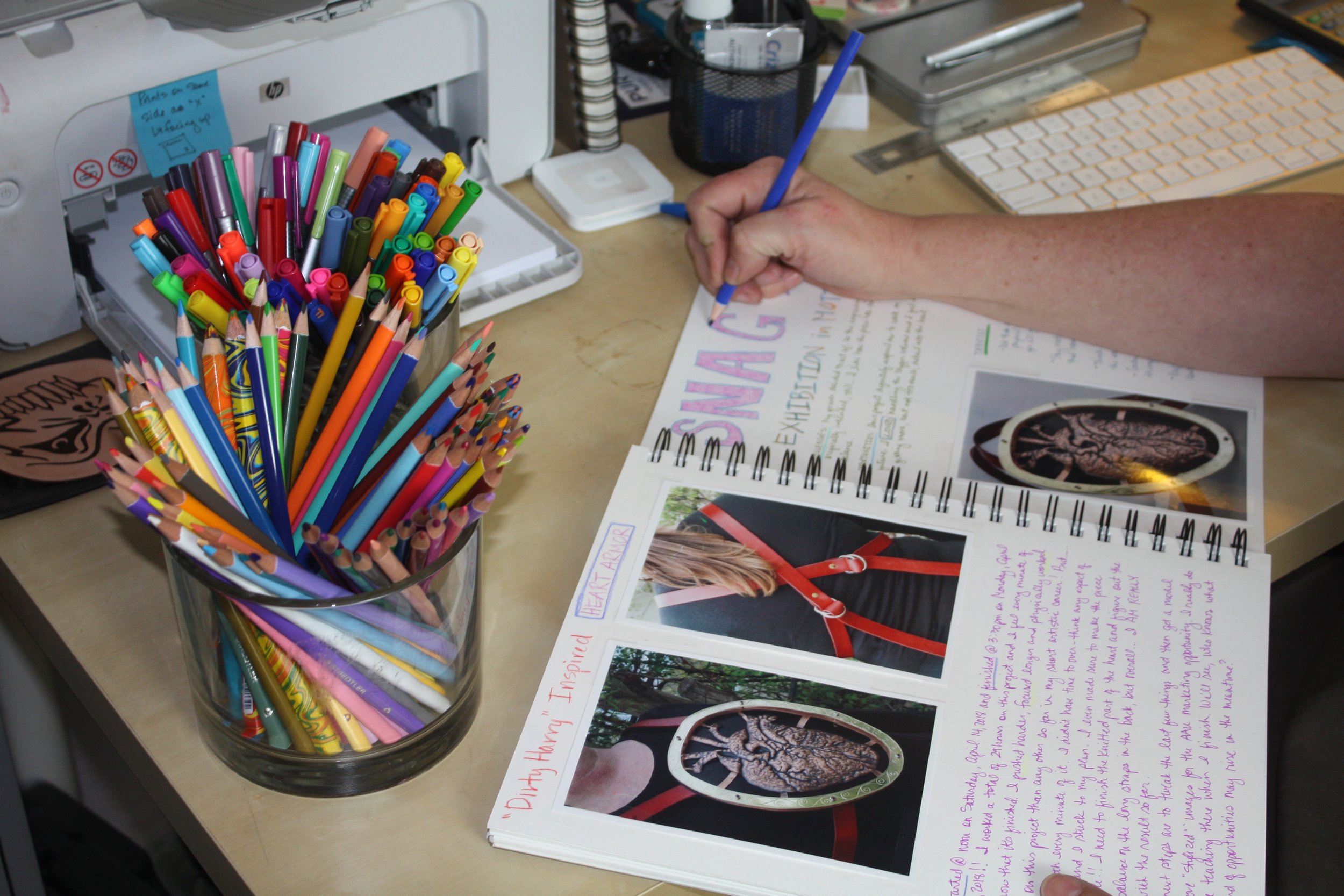 A desk with a clear container holding various colorful markers, a patient's note in a notebook with handwritten text and images of brain scans, a computer keyboard, a pen, and office supplies.