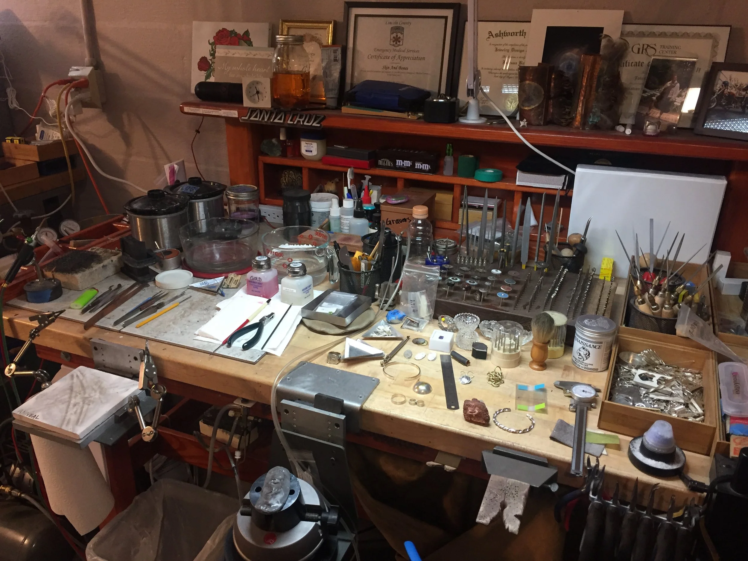Messy workbench with tools, jewelry, and various objects on the table and shelf, surrounded by certificates and framed artwork.