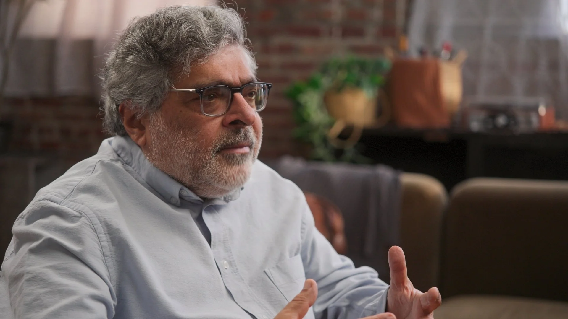 An older man with glasses, gray hair, wearing a dark blazer and a white shirt, sitting indoors and talking, with blurred interior background.