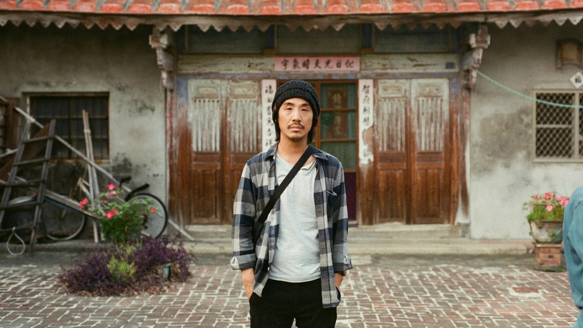A young man with long dark hair, wearing a black knit beanie, a gray t-shirt, a plaid shirt, and black pants, stands in front of an old wooden building with Chinese characters on a sign above the door.