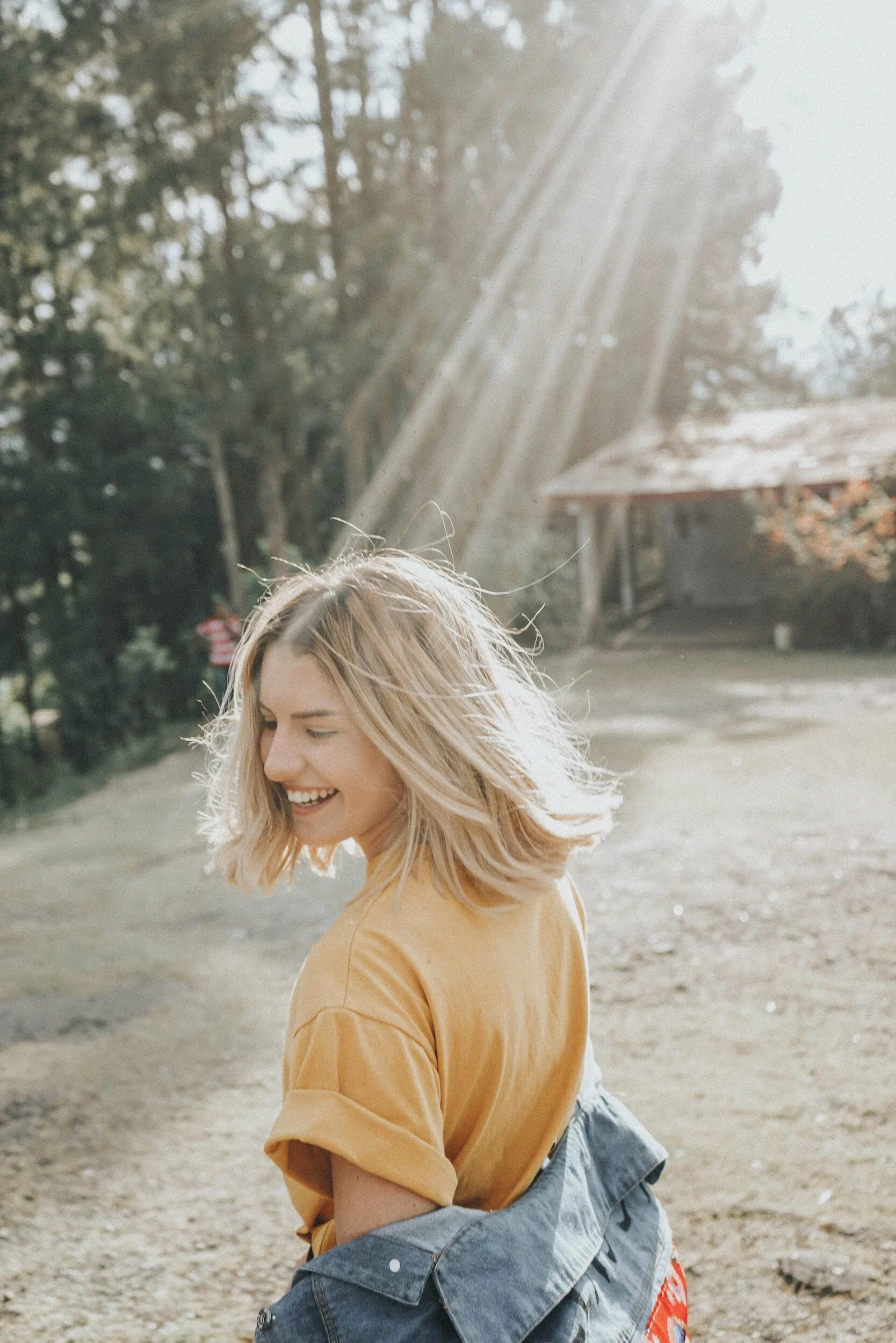 A smiling person with blonde hair wearing a yellow shirt and a denim jacket in a sunny outdoor setting.