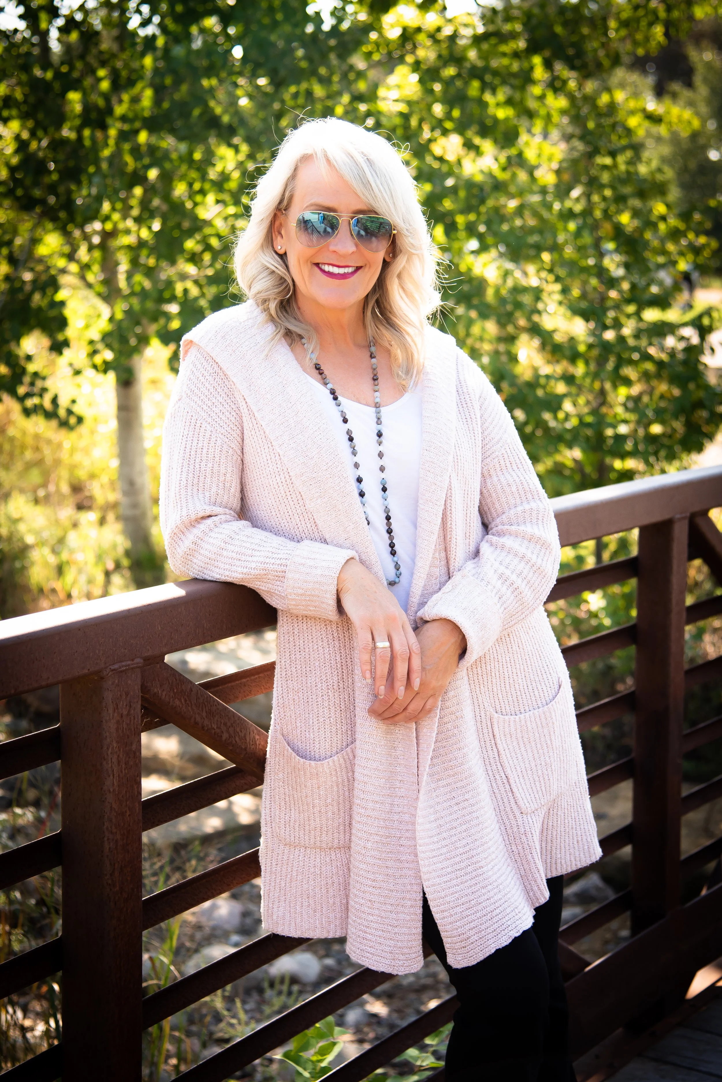 Woman with sunglasses and long blonde hair standing on a wooden bridge, wearing a light pink cardigan and a necklace, with trees in the background.