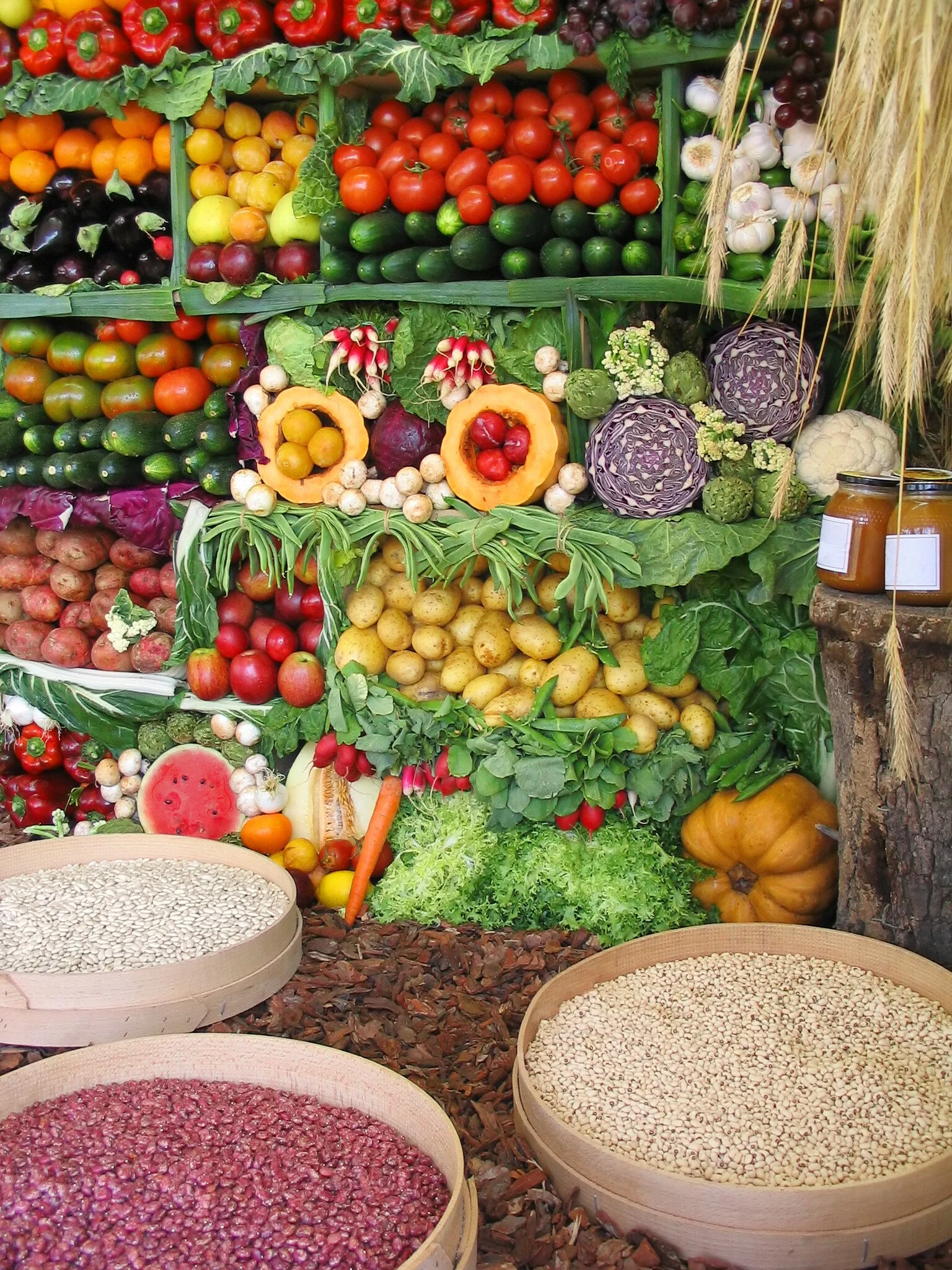 Colorful display of fresh vegetables and fruits at a market, including tomatoes, cucumbers, eggplants, cabbages, pumpkins, radishes, lettuce, carrots, and pumpkins, with some jars of jam and containers of seeds in the foreground.